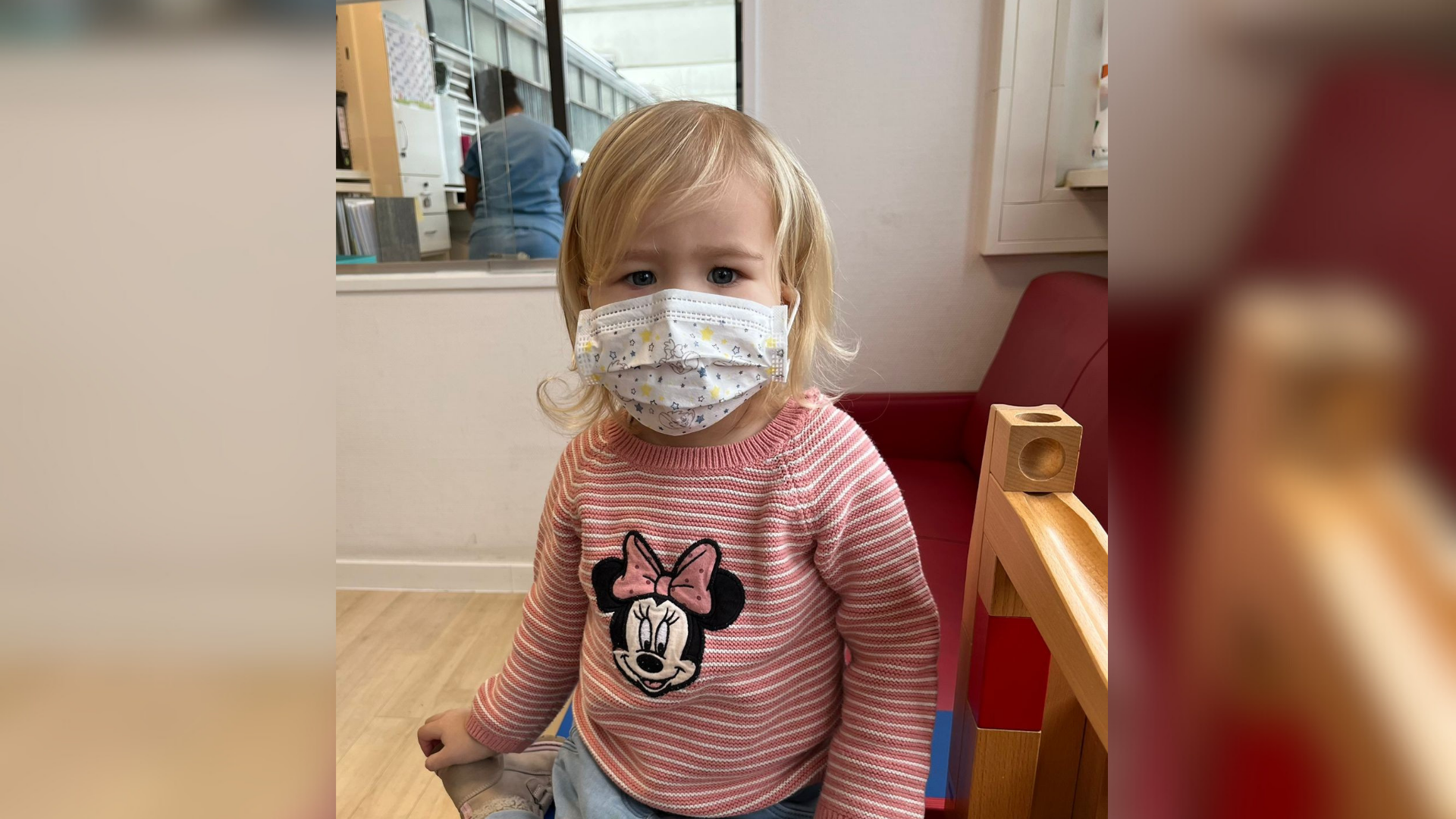 Ein Mädchen sitzt mit Mundschutz und einem Minnie-Maus-Oberteil auf einer Spielmatte.