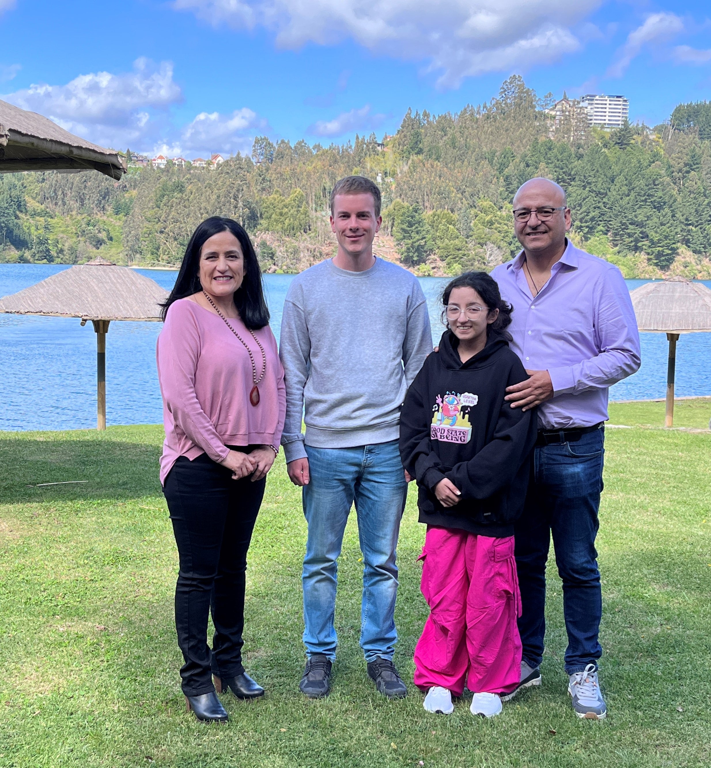 Vier Personen stehen nebeneinander auf einer Rasenfläche mit Blick auf einen See. Im Hintergrund sind runde Strohdächer, Wasser, bewaldete Hügel und ein Gebäude auf einem Hang zu sehen. Die Frau links trägt ein rosa Oberteil und dunkle Hose, der Mann rechts ein hellblaues Hemd und hat den Arm um das Mädchen neben sich gelegt, das eine schwarze Kapuzenjacke und eine auffällig pinke Hose trägt. Der junge Mann in der Mitte trägt einen grauen Pullover und Jeans. Alle blicken in die Kamera.