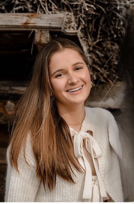 Ein Portrait einer lächelnden jungen Frau. Sie trägt helle Kleidung und steht vor einem neutralen Hintergrund. 