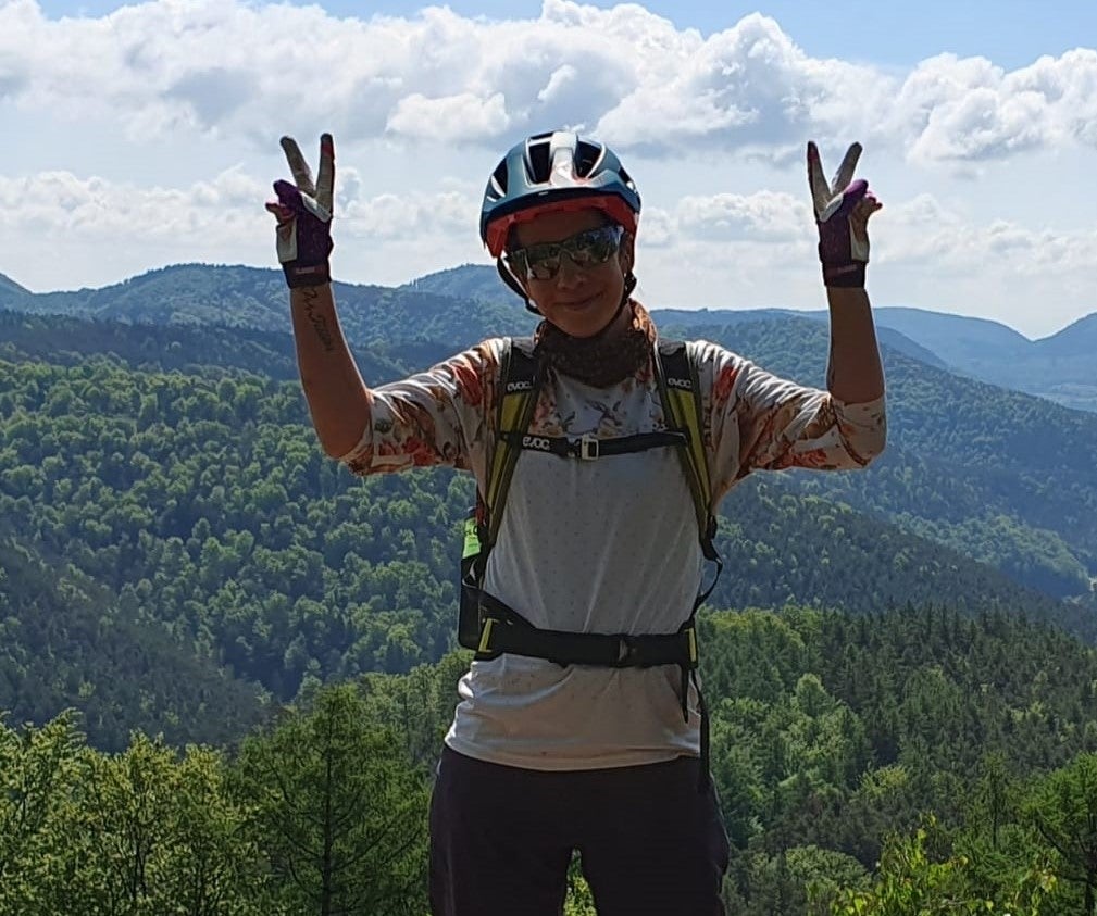 Eine Frau steht mit Fahrradhelm und Rennbrille auf einem Berg und hält Peace-Zeichen in die Luft.