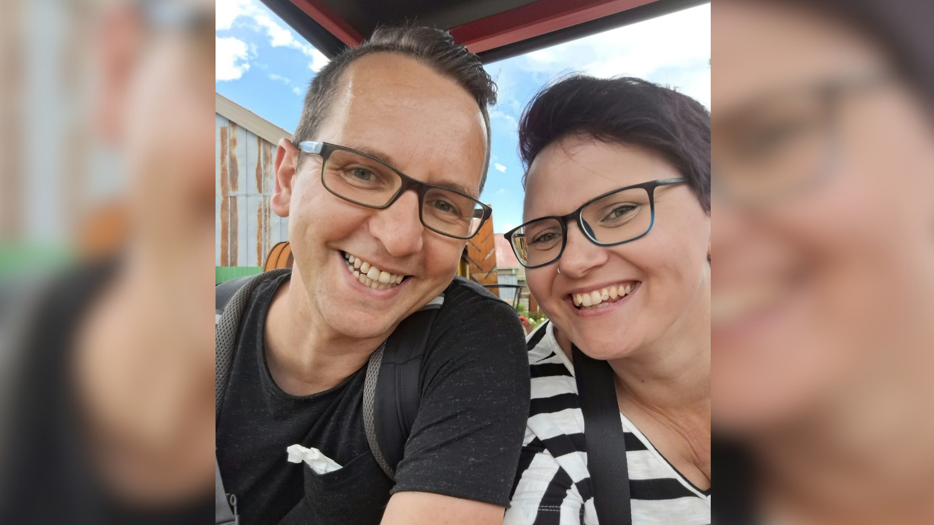 Ein Mann trägt ein schwarzes T-Shirt. Eine Frau trägt ein schwarz-weiß gestreiftes T-Shirt. Beide tragen Brille und lächeln in die Kamera. 