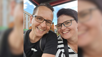 Ein Mann trägt ein schwarzes T-Shirt. Eine Frau trägt ein schwarz-weiß gestreiftes T-Shirt. Beide tragen Brille und lächeln in die Kamera. 