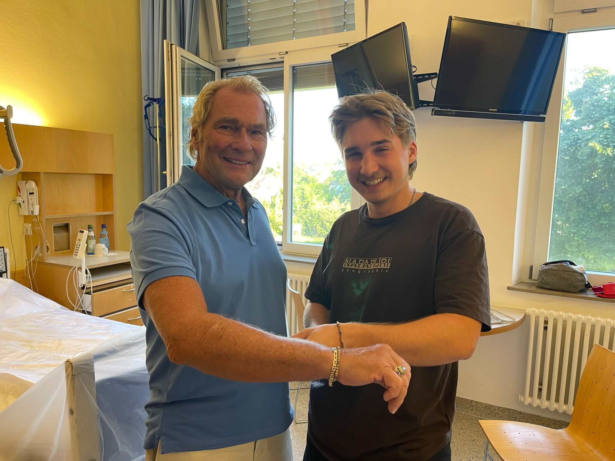 Zwei Menschen stehen in einem Krankenhauszimmer und halten ein Armband in die Kamera