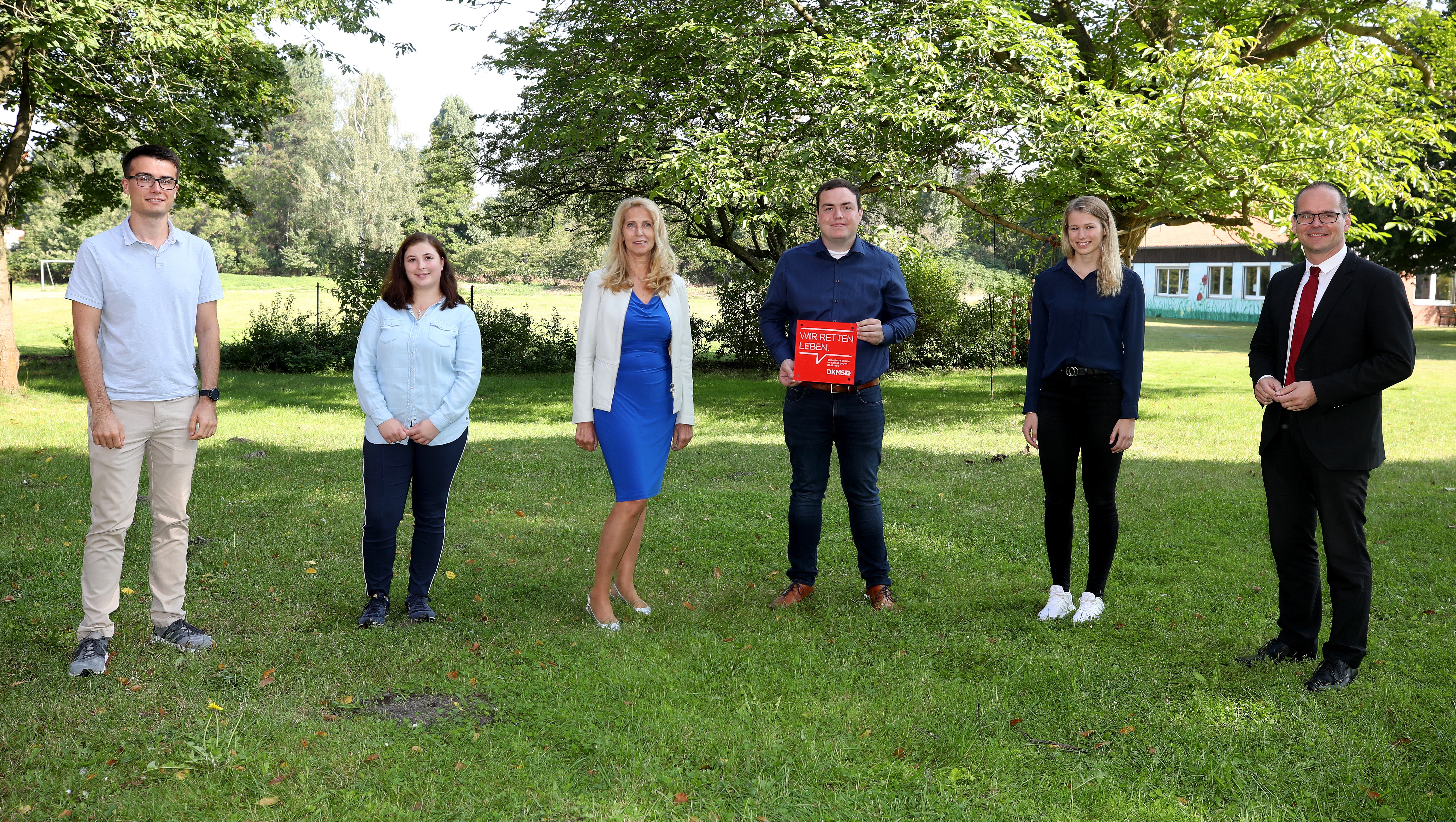 6 Menschen posieren auf einer Wiese für ein Foto, einer hält eine Tafel mit der Aufschrift: "Wir retten Leben" in der Hand