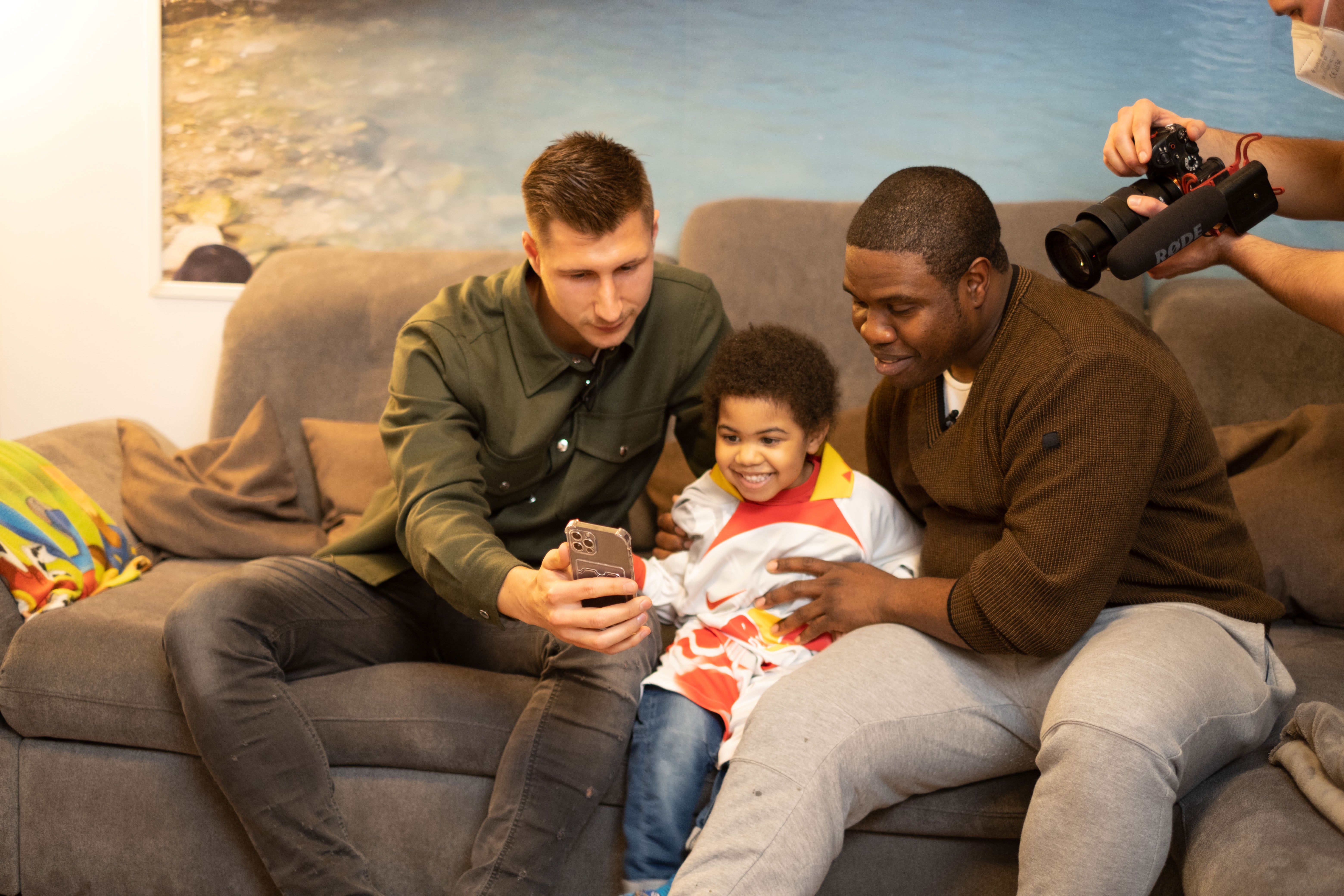 zwei Erwachsene sitzen mit einem Kind auf einem Sofa und gucken auf ein Handy