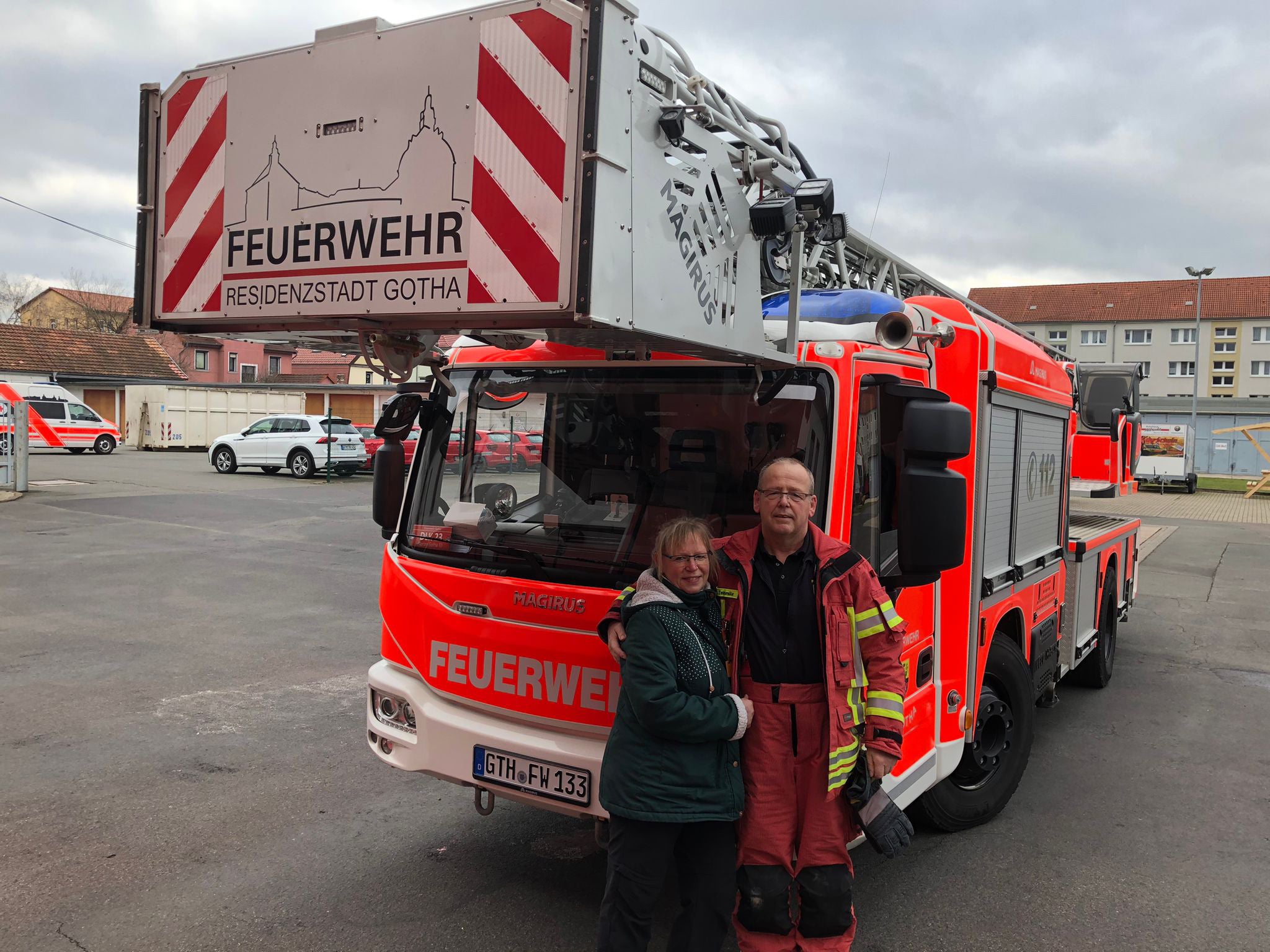 Zwei Personen stehen lächelnd vor einem großen roten Feuerwehrauto mit der Aufschrift „Feuerwehr Residenzstadt Gotha“. Eine Person trägt eine Feuerwehruniform, die andere eine grüne Jacke. Im Hintergrund sind weitere Einsatzfahrzeuge und Gebäude zu sehen.