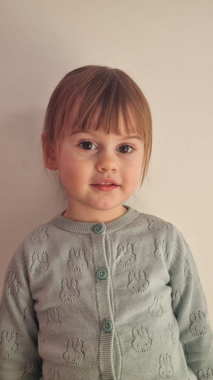 Ein kleines Mädchen mit braunen Haaren lächelt in die Kamera. Sie trägt eine graue Strickjacke auf der Häschen zu sehen sind.