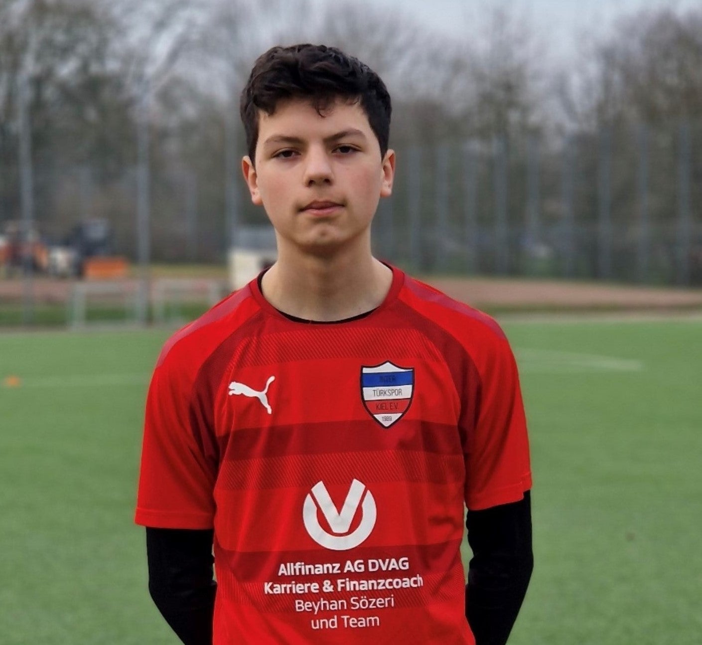 Ein Junge in einem roten Trikot, im Hintergrund sieht man einen Fußballplatz