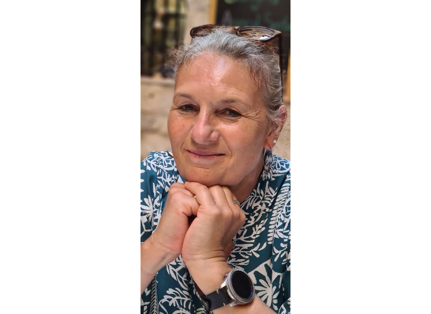 Eine Frau mit ergrautem Haar und einer auf dem Haar aufliegenden Brille stützt ihr Kinn auf den zu Fäusten geschlossenen Händen und lächelt.