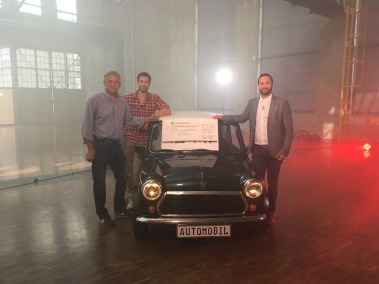 Eine Gruppe von Männern steht neben einem Auto. Auf  der Frontscheibe liegt ein großer Scheck.