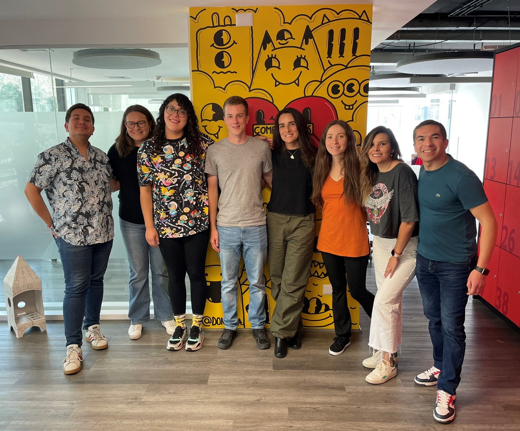 Acht Personen stehen nebeneinander in einem modernen Büroraum und lächeln in die Kamera. Hinter ihnen befindet sich eine gelbe Wand mit schwarzer Comic-Grafik und roten Akzenten. Alle Personen tragen Freizeitkleidung in unterschiedlichen Farben. Der Raum hat Holzfußboden, Glaswände und eine moderne Decke mit offenen Kabelkanälen.