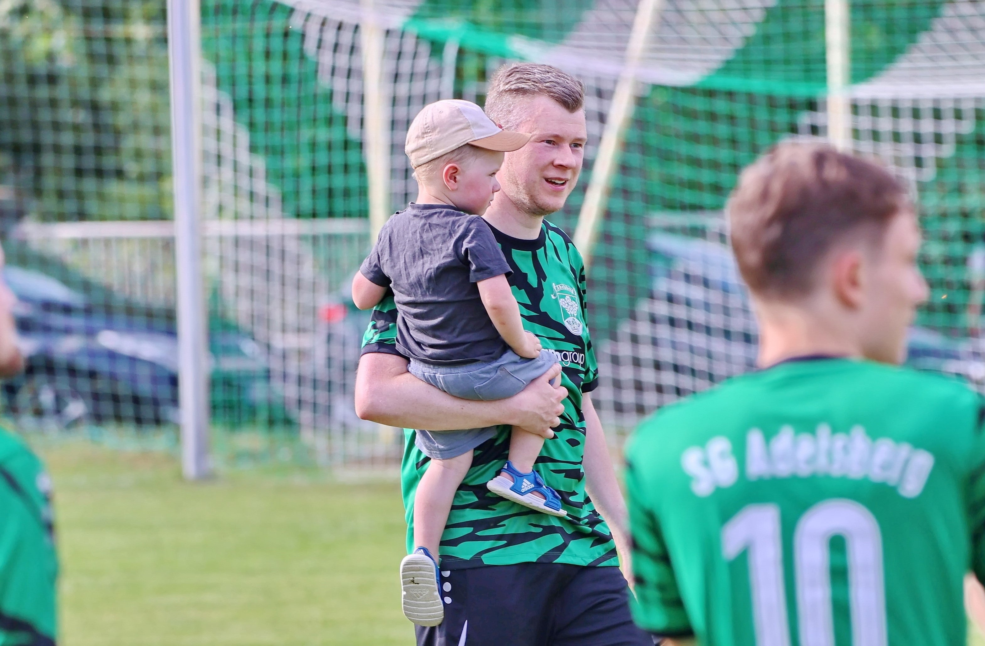 ein Mann steht auf einem Rasen, hinter ihm ein Fußballtor und er hält ein Kind auf dem Arm