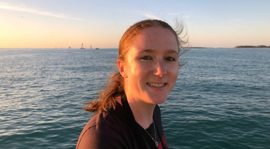 Eine Frau lächelt in die Kamera. Im Hintergrund sieht man Meer.