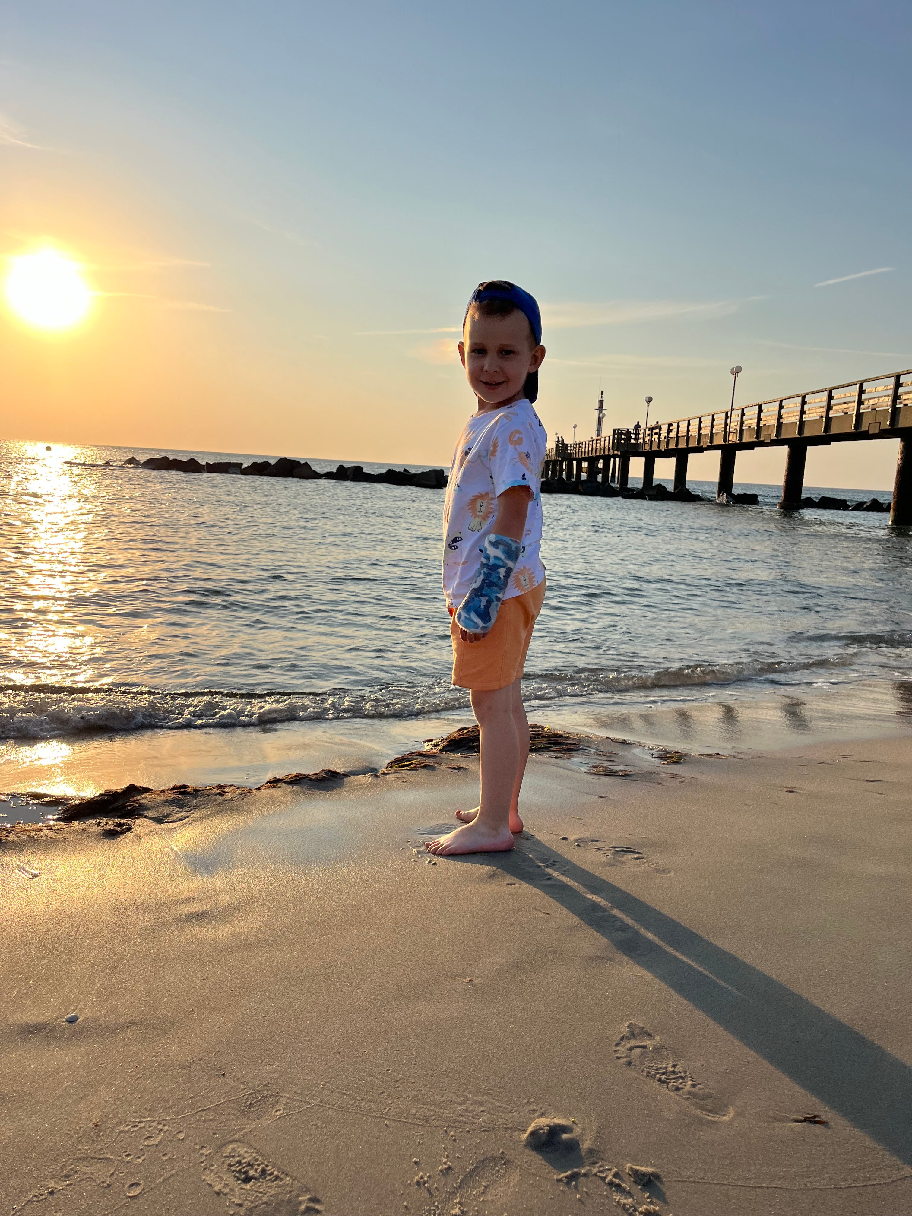 Ein Kind steht am Strand. Im Hintergrund sieht man das Meer, die untergehende Sonne und einen Steg 