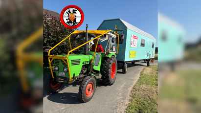 Ein Mann fährt einen grünen Traktor, der einen türkisfarbenen Bauwagen zieht. 
