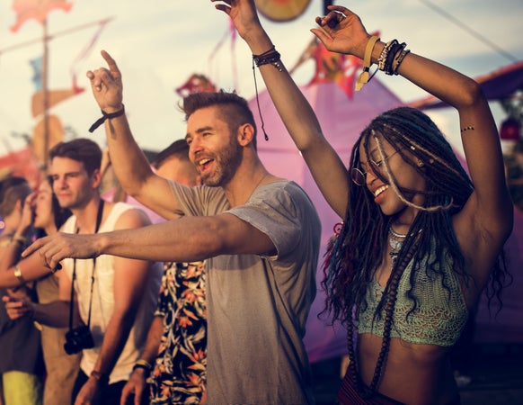 Mehrere junge Erwachsene tanzen und feiern bei einem Open-Air-Festival. Im Vordergrund hebt eine Frau mit langen geflochtenen Haaren und ein Mann mit Bart und gestreiftem T-Shirt fröhlich die Arme in die Luft. Die Stimmung ist ausgelassen, die Sonne scheint, und im Hintergrund sind bunte Dekorationen und weitere Festivalbesucher\:innen zu sehen.
