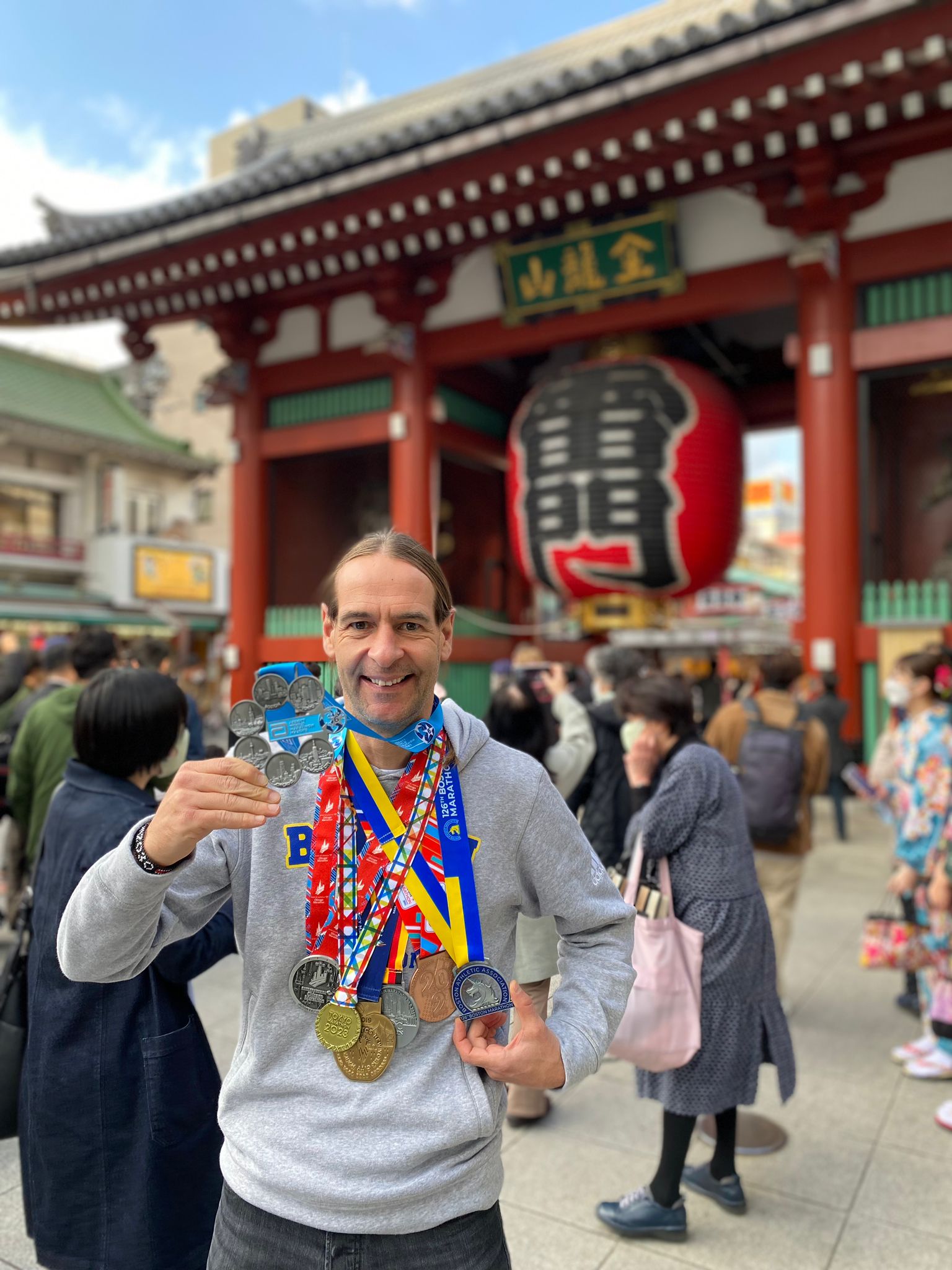 Ein Mann steht vor dem Eingang eines Tempels und hält mehrere Medaillen in der Hand. Er trägt weitere Medaillen um den Hals. Im Hintergrund sind viele Menschen vor dem Tempel zu sehen.