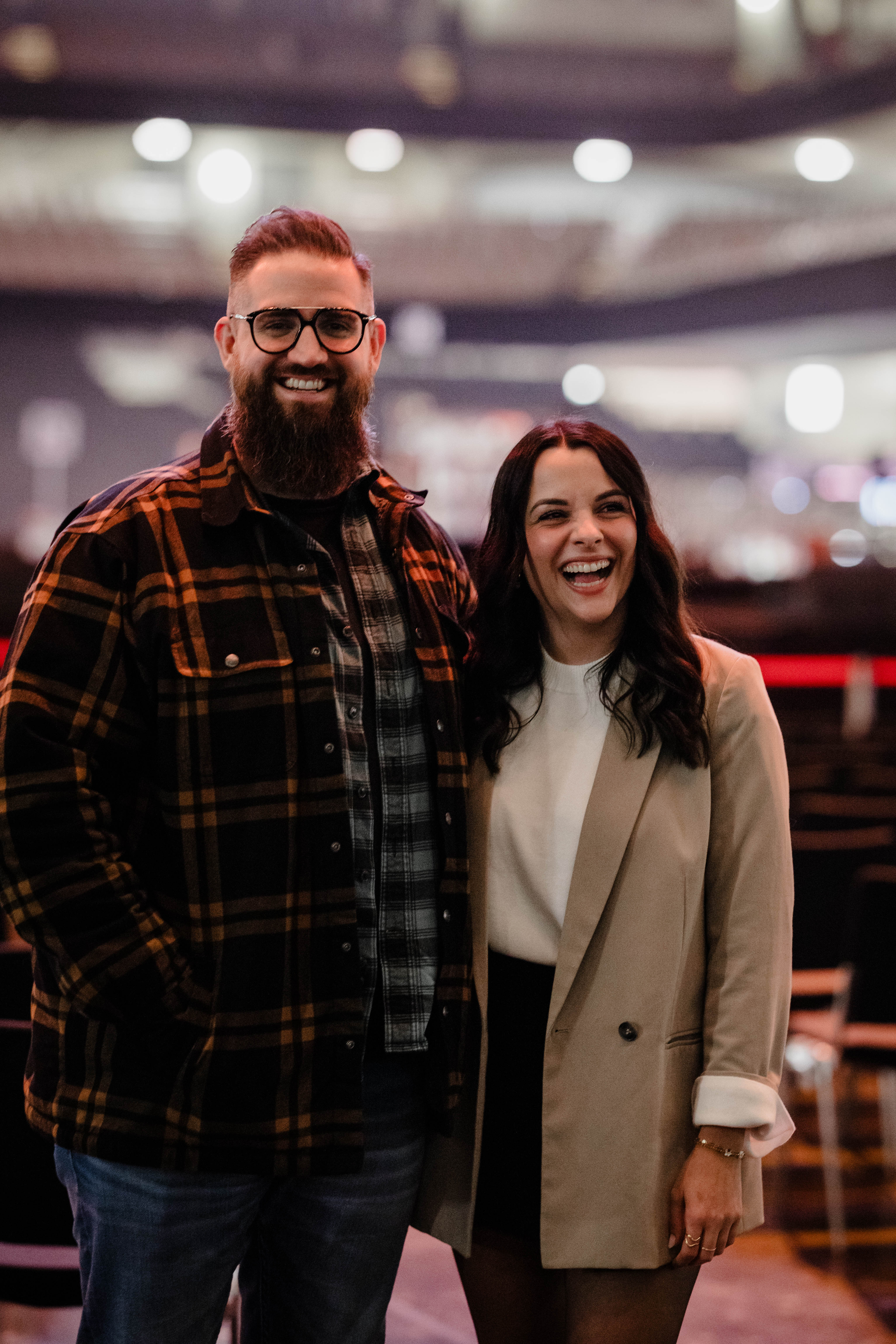 Ein Mann im roter kartierter Jacke und eine Frau im beigen Mantel lächeln in die Kamera
