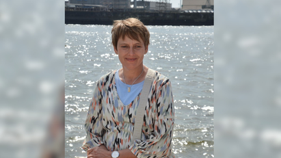 Eine Frau sitzt an einem Gewässer. Im Hintergrund ist ein Hafen zu sehen.