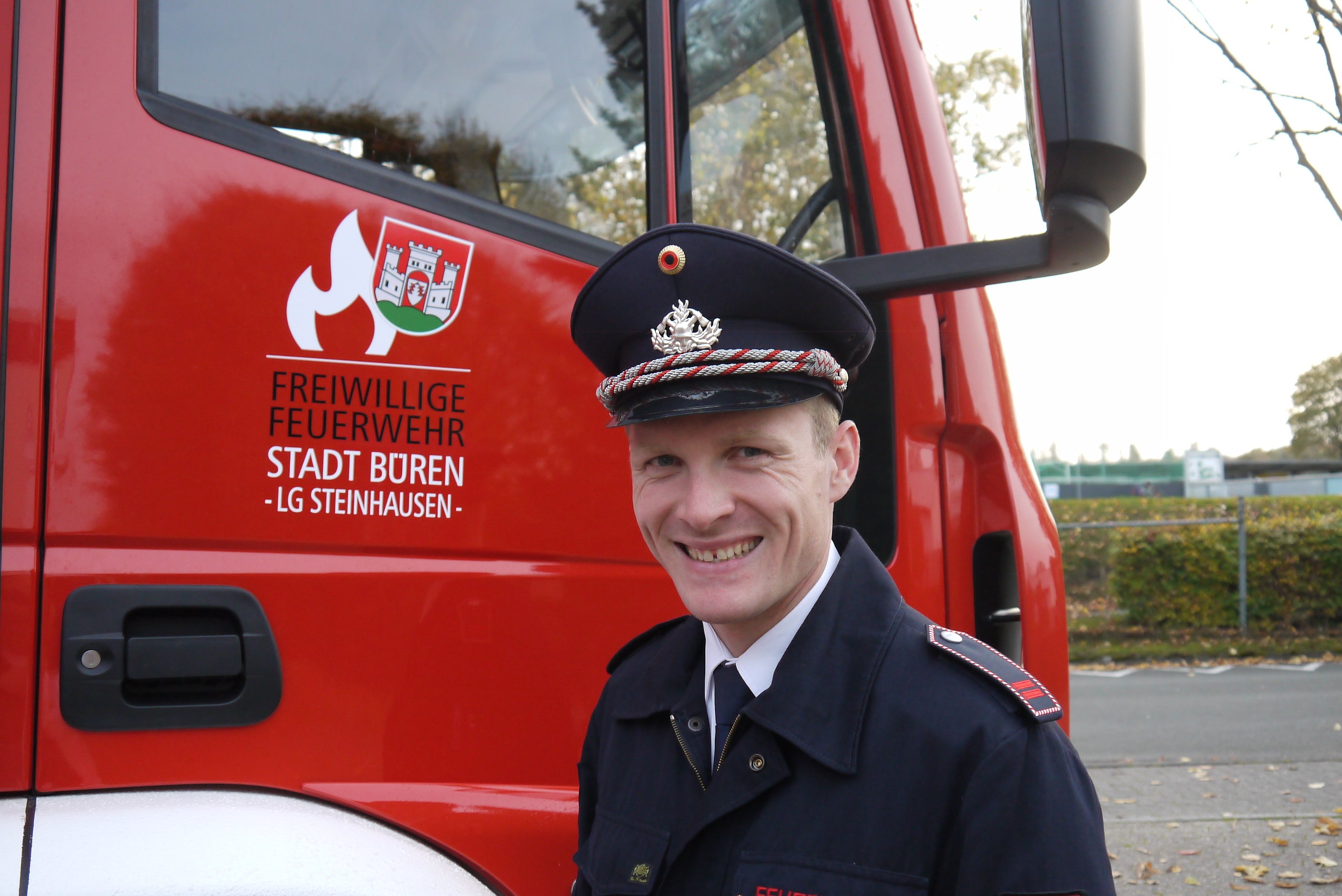 Ein Feuerwehrmann in Uniform mit Schirmmütze steht lächelnd vor einem roten Einsatzfahrzeug der Freiwilligen Feuerwehr Stadt Büren, Löschgruppe Steinhausen. Auf dem Fahrzeug ist das Feuerwehr-Logo mit Flamme und Stadtwappen zu sehen.