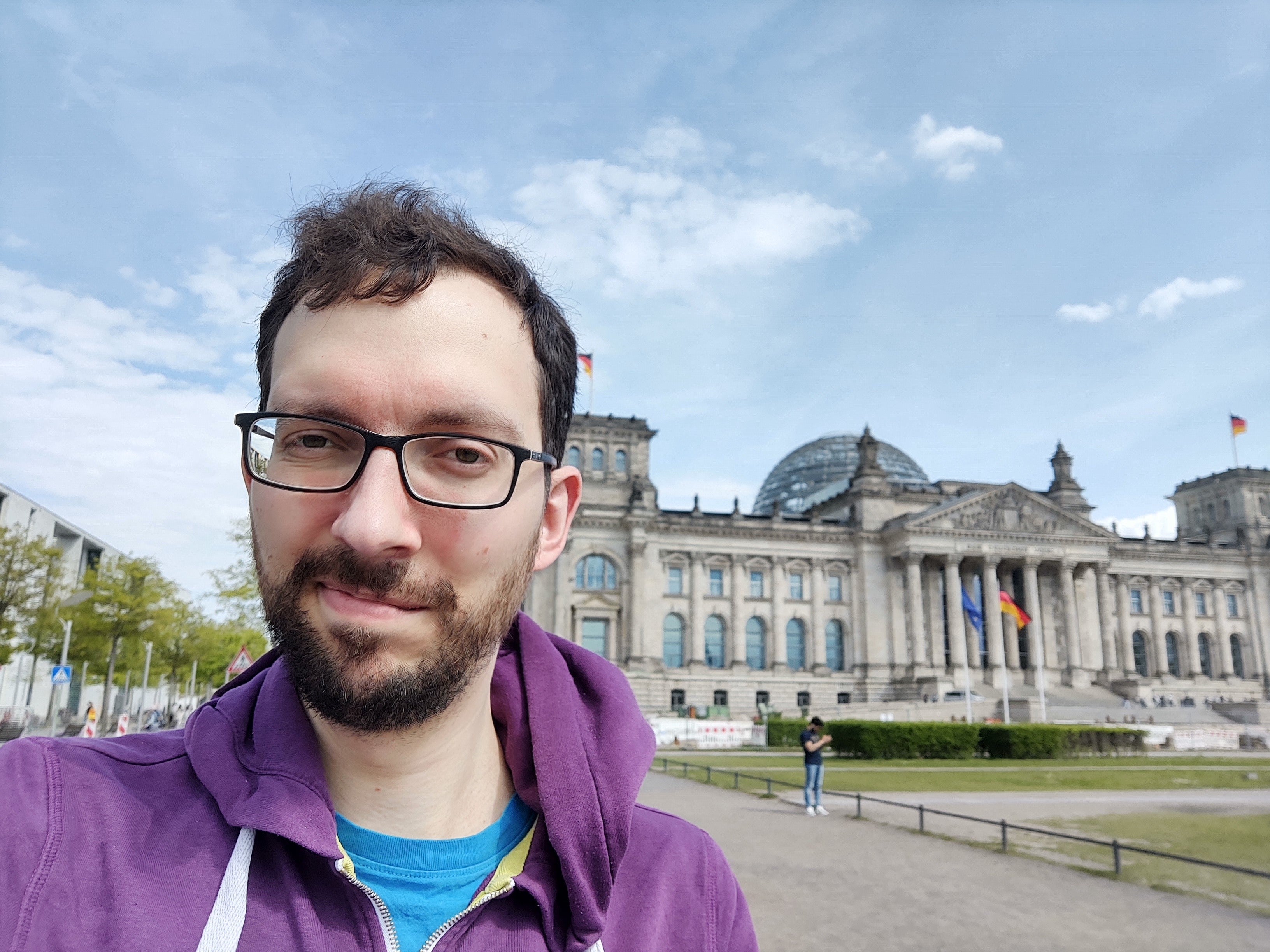 Ein Mann mit Brille und Bart trägt eine lila Kapuzenjacke über einem blauen T-Shirt und macht ein Selfie vor dem Reichstagsgebäude in Berlin. Im Hintergrund sind die Reichstagskuppel, deutsche Flaggen und der Vorplatz mit wenigen Personen zu sehen. Der Himmel ist leicht bewölkt.