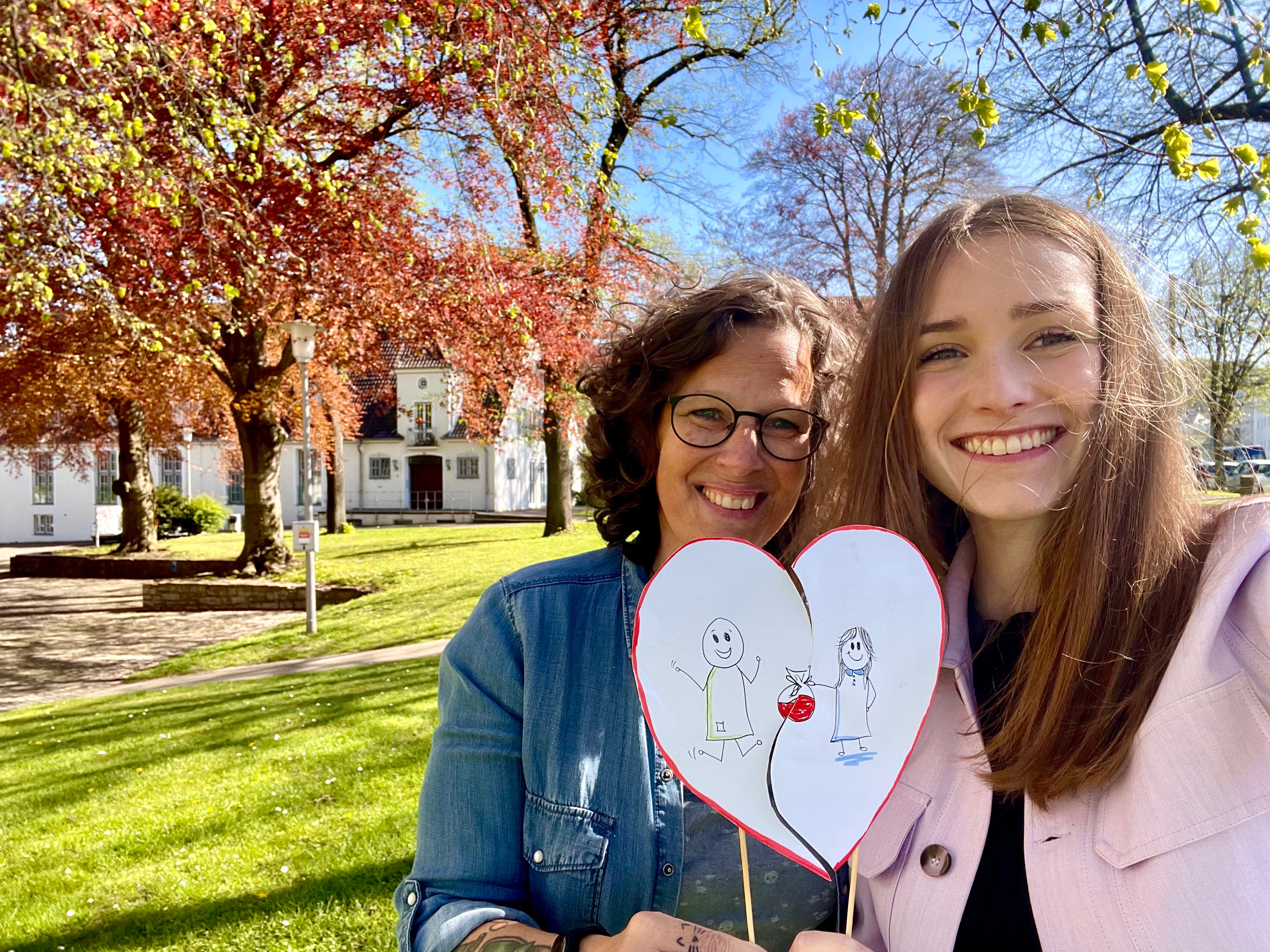 Zwei Frauen stehen auf einer Wiese im Sonnenschein und lächeln in die Kamera. Sie halten gemeinsam ein herzförmiges Schild mit einer Kinderzeichnung, die zwei Personen mit einem verbundenen Herzen zeigt. Im Hintergrund stehen blühende Bäume und ein weißes Gebäude mit rotem Dach.