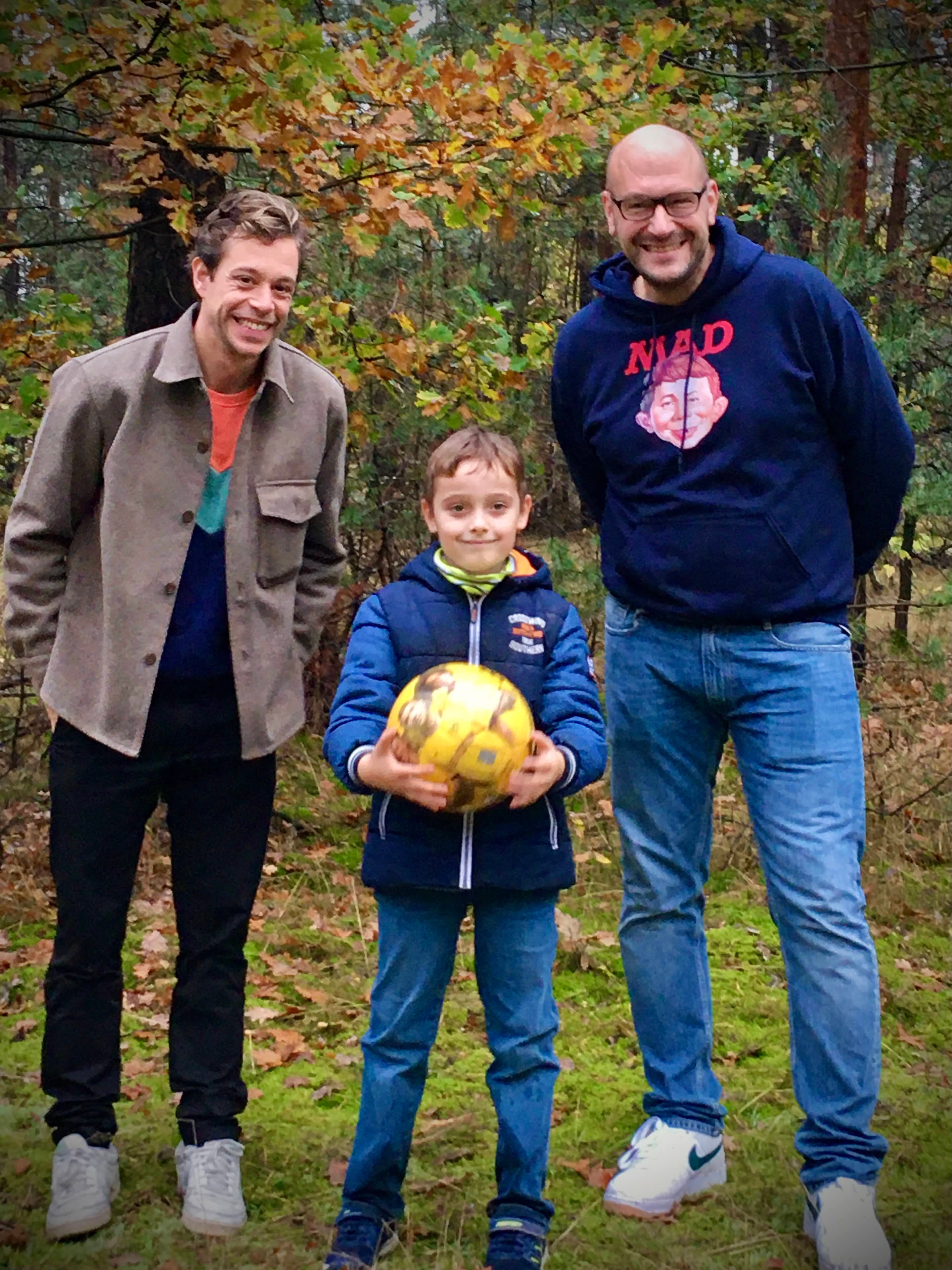 zwei Männern stehen vor einem Wald mit einem Jungen, der einen gelben Ball hält