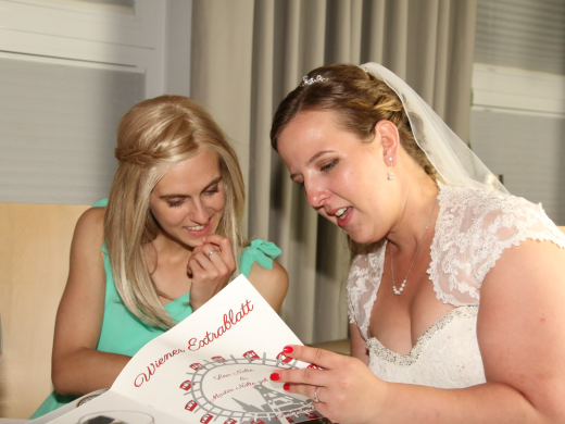 Eine Frau in einem Hochzeitskleid schaut in ein Buch, daneben steht eine andere Frau die mit in das Buch schaut 