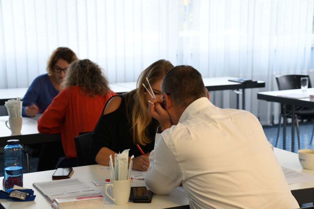 Verschiedene Personen sitzen sich an Tischen gegenüber. Ein Mann im Vordergrund hält mehrere Wattestäbchen in der Hand. 