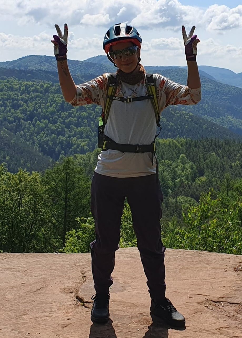Ilona steht in Fahrradkleidung vor einer Bergkulisse. Sie lächelt in die Kamera und hält beide Hände zu Peace-Zeichen hoch.