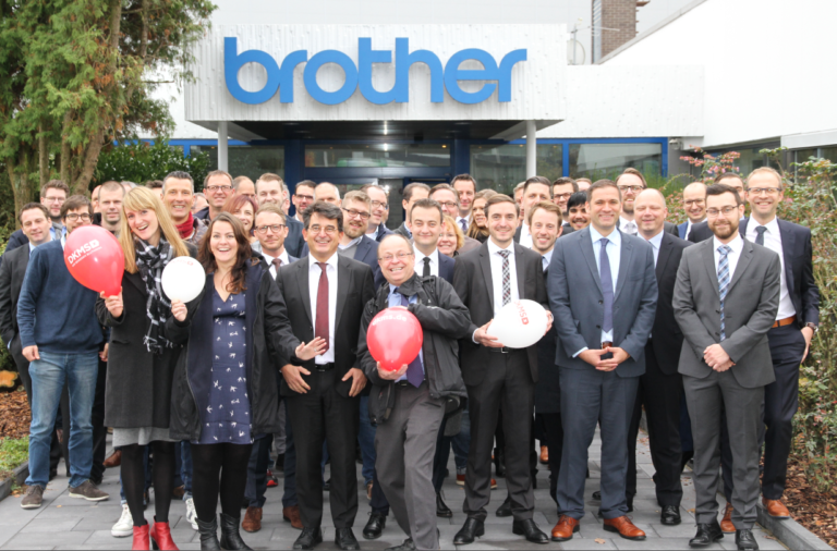 Eine Gruppe von Menschen posiert vor einem Gebäude. Auf dem Gebäude ist ein Logo der Firma brother. 