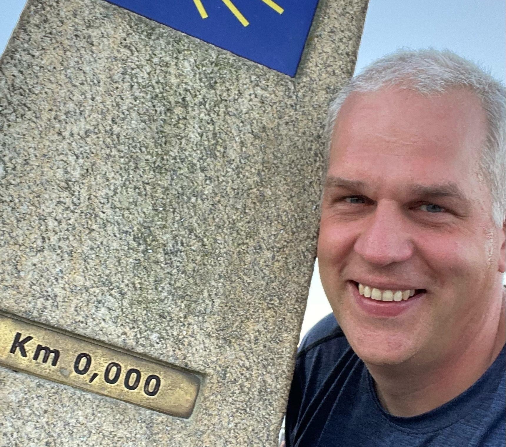 Markus lehnt an einem Pilgerwegmarker und lächelt in die Kamera.