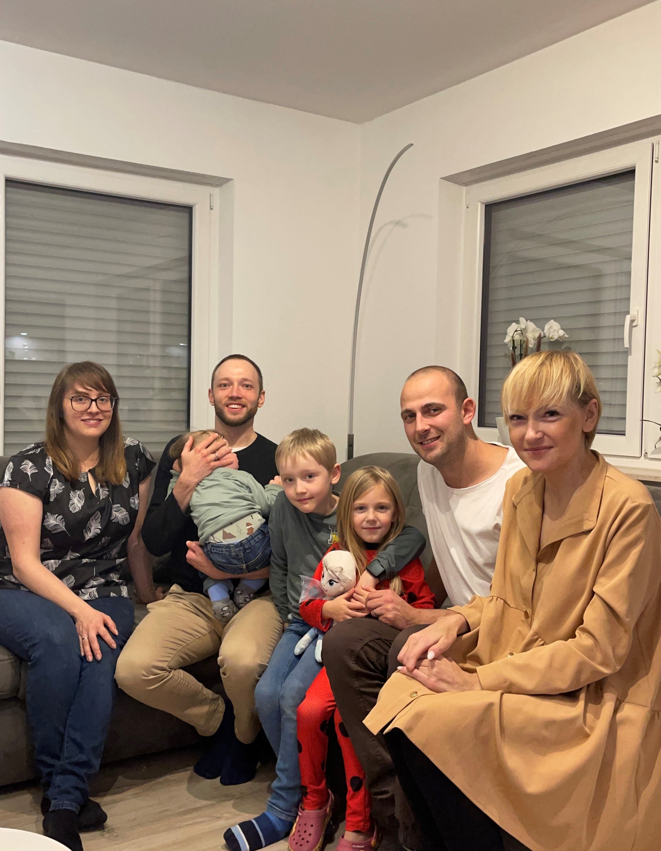 Gruppenfoto in einem Wohnzimmer: Sieben Personen sitzen eng beieinander auf einem Sofa und einem Hocker. Zu sehen sind drei Erwachsene, zwei Männer mit kurzen Haaren, eine Frau mit blonder Kurzhaarfrisur, daneben eine weitere Frau mit Brille, ein Baby auf dem Arm eines Mannes sowie zwei Kinder in der Mitte, ein Junge und ein Mädchen mit Kuscheltier. Alle lächeln in die Kamera.