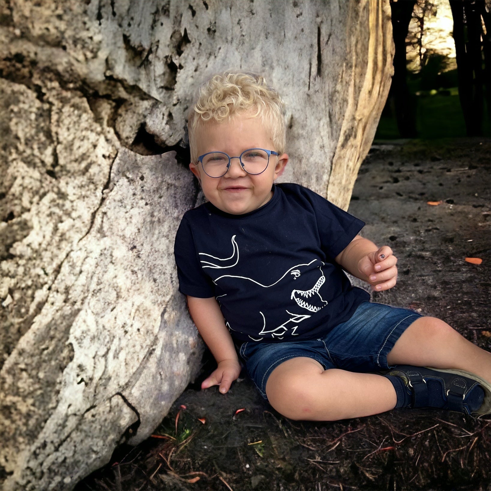 kleines Kind mit blonden Haaren und Brille sitzt auf dem Boden und lehnt an einen Baum