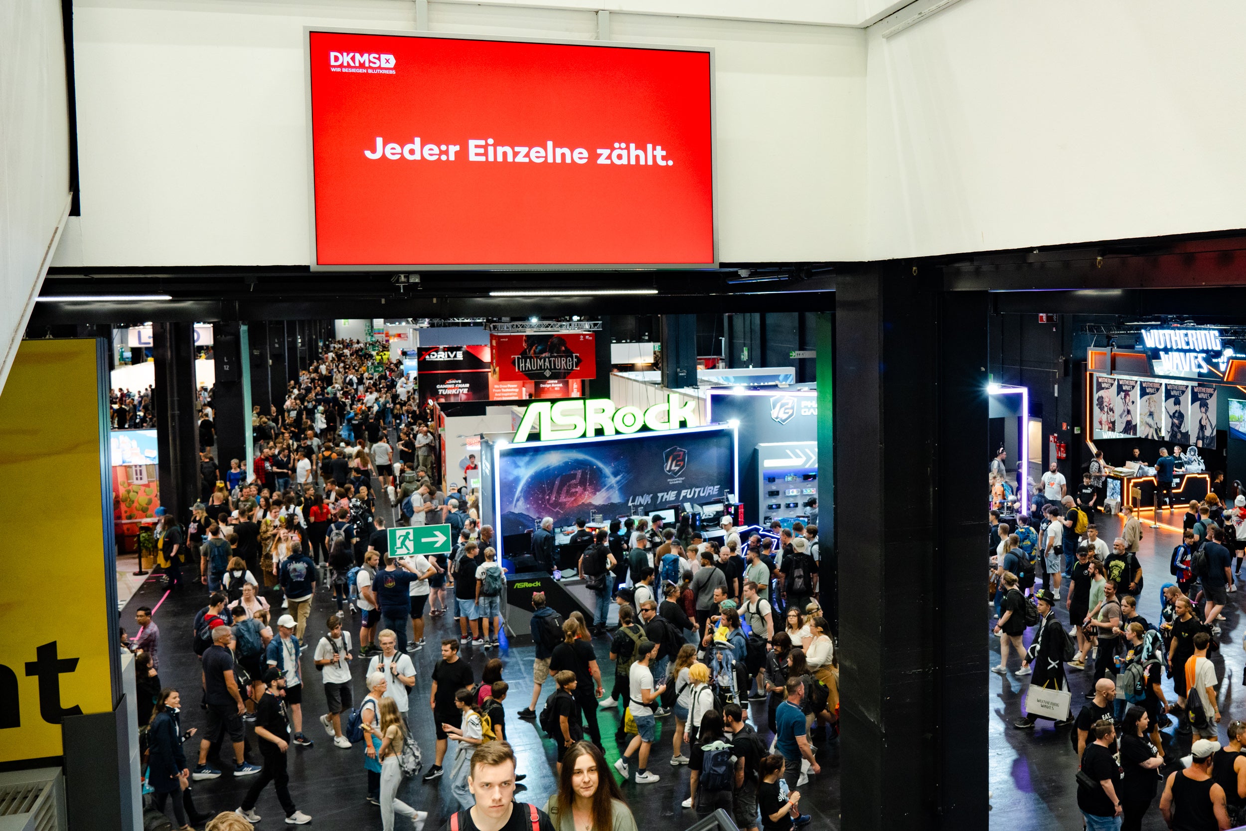 Das Bild zeigt eine Messehalle mit zahlreichen Besucherinnen und Besuchern. Im oberen Bereich hängt ein großer roter Bildschirm mit weißem Text: „DKMS – Jeder: Einzelne zählt.“ Darunter befinden sich verschiedene Messestände, unter anderem mit der Aufschrift „ASROCK“. Die Halle ist gut gefüllt und es herrscht reger Betrieb.
