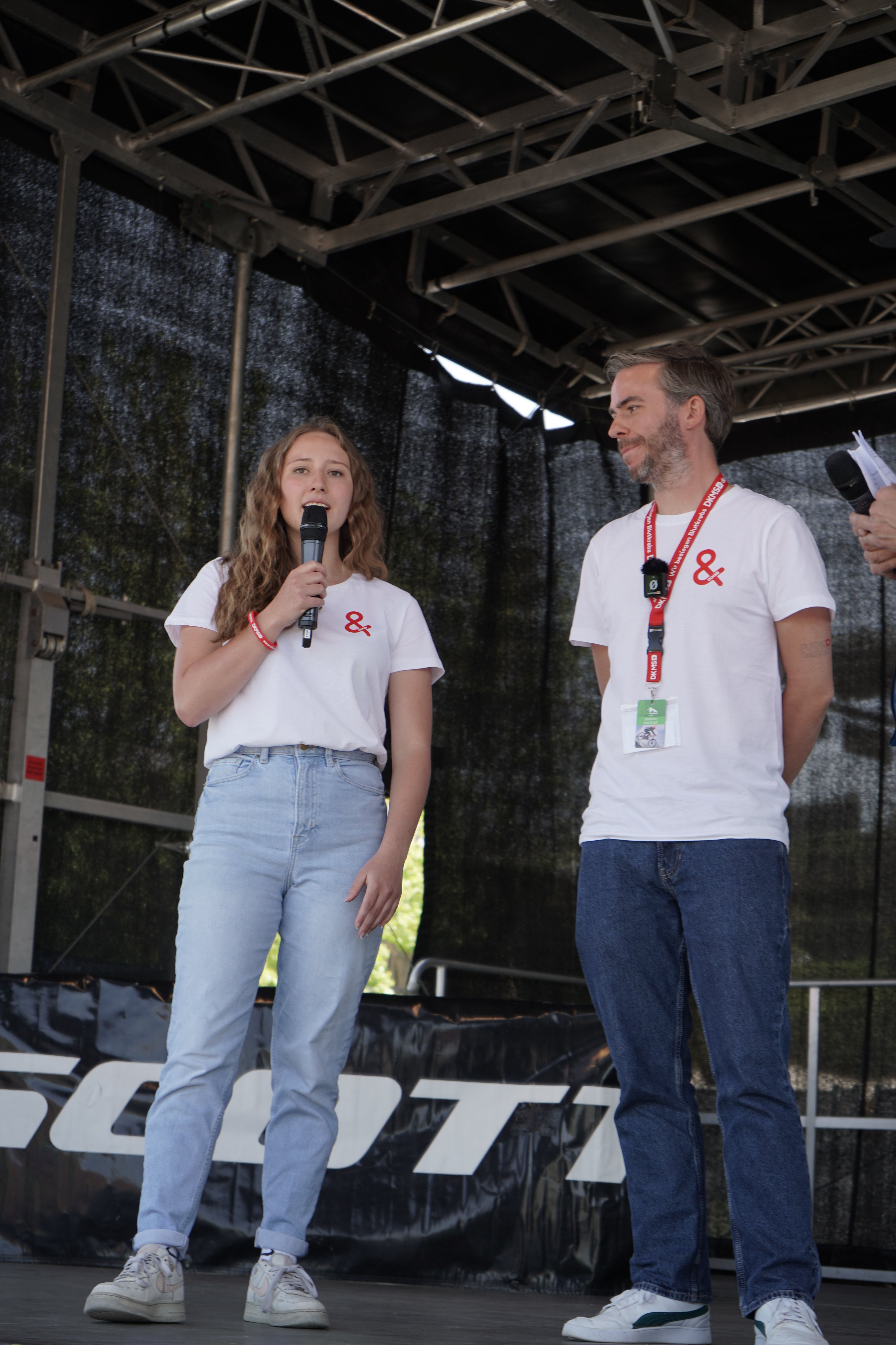 Eine Frau und ein Mann stehen auf einer Bühne unter einem Metalldach. Beide tragen weiße T-Shirts mit rotem &-Symbol sowie blaue Jeans. Die Frau hält ein Mikrofon. Beide tragen Veranstaltungsbändchen und Ausweise um den Hals. Im Hintergrund ist ein schwarzer Bühnenvorhang zu sehen.