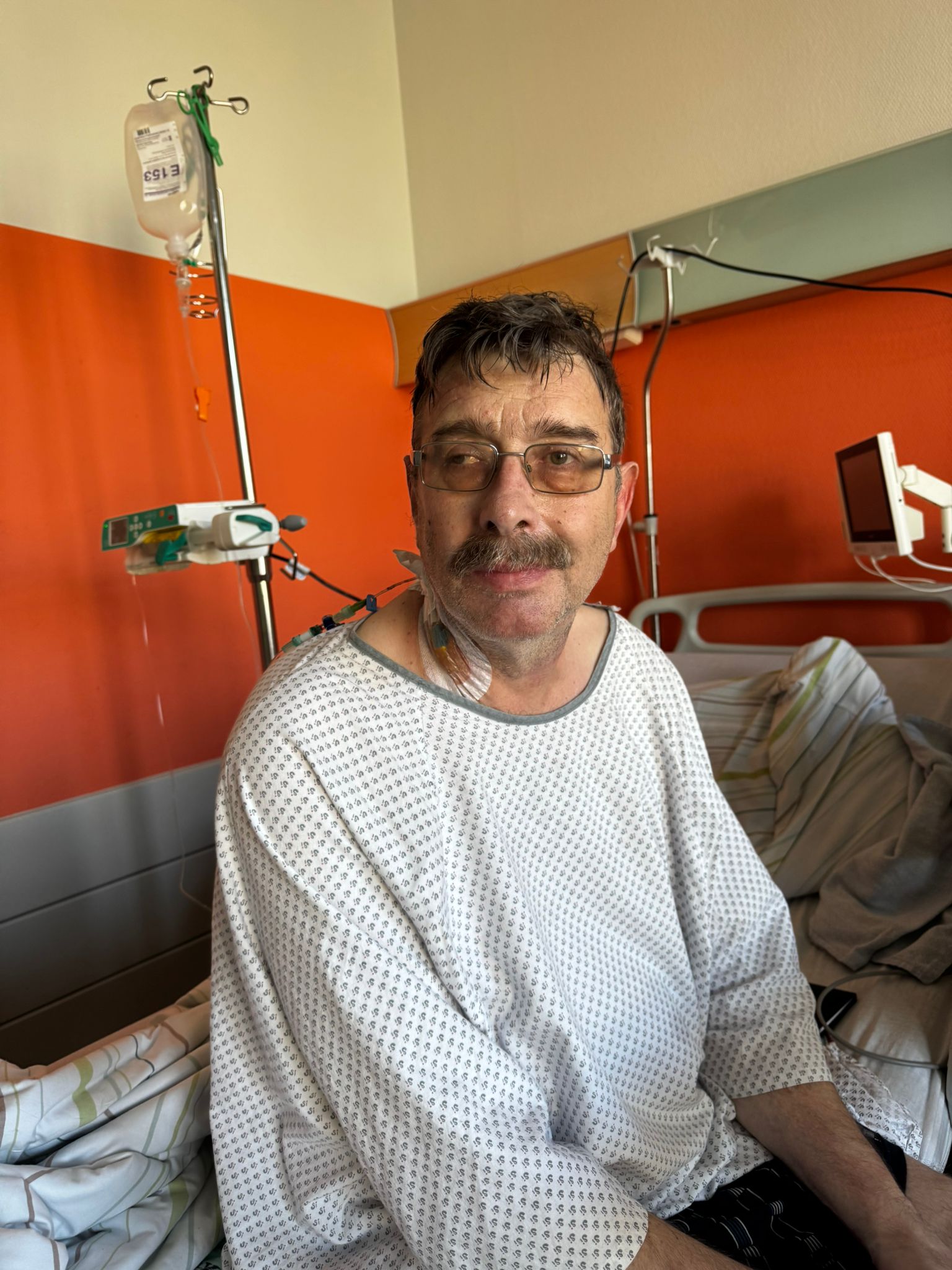 Mann mit Brille sitzt in Krankenhausrobe auf Krankenhausbett