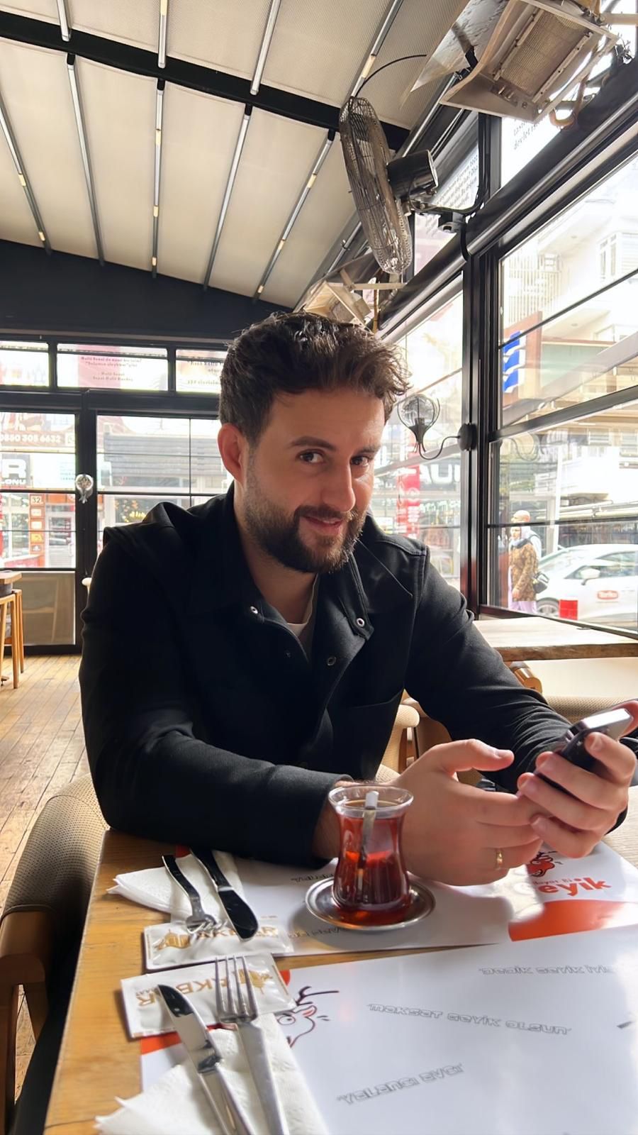 DKMS-Patient Harun sitzt in einem Cafe und lächelt. In seiner Hand hält er ein Handy. 