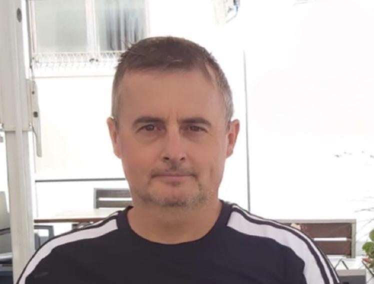 Ein Mann mit kurzem Haar und leichtem Bart sitzt draußen vor einem hellen Hintergrund. Er trägt ein dunkelblaues oder schwarzes T-Shirt mit weißen Streifen an den Schultern. Im Hintergrund sind Fenster, ein Tisch und helle Strukturen zu sehen.