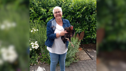 Eine ältere Frau steht vor einem grünen Bett und hält ein braun gefiedertes Huhn.