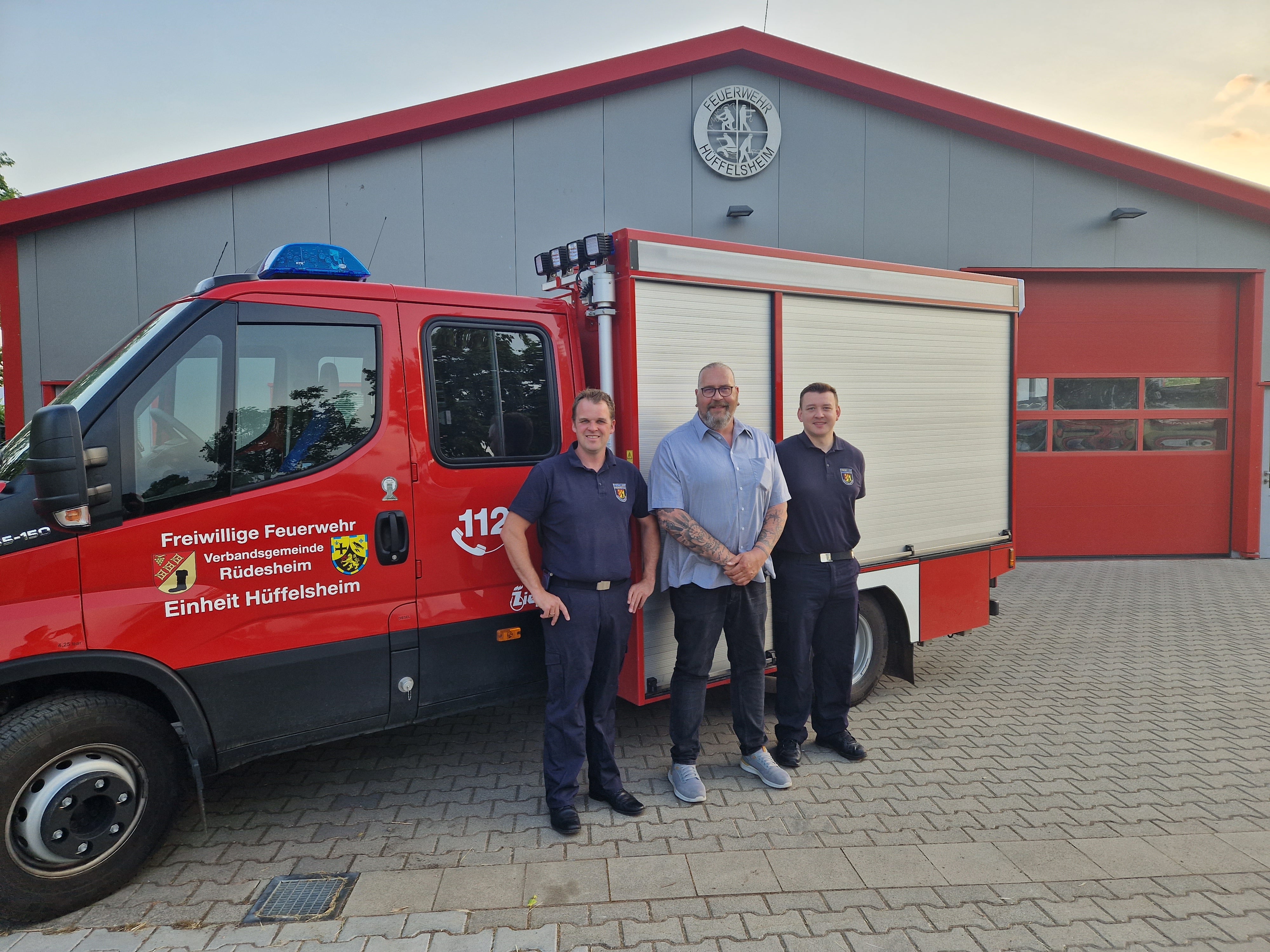 Drei Männer stehen vor einem roten Einsatzfahrzeug der Freiwilligen Feuerwehr Rüdesheim, Einheit Hüffelsheim. Zwei der Männer tragen Feuerwehruniform, der dritte Mann steht in der Mitte in Freizeitkleidung. Im Hintergrund ist ein graues Feuerwehrgebäude mit rotem Tor und Wappen über dem Eingang zu sehen. Der Boden ist gepflastert.
