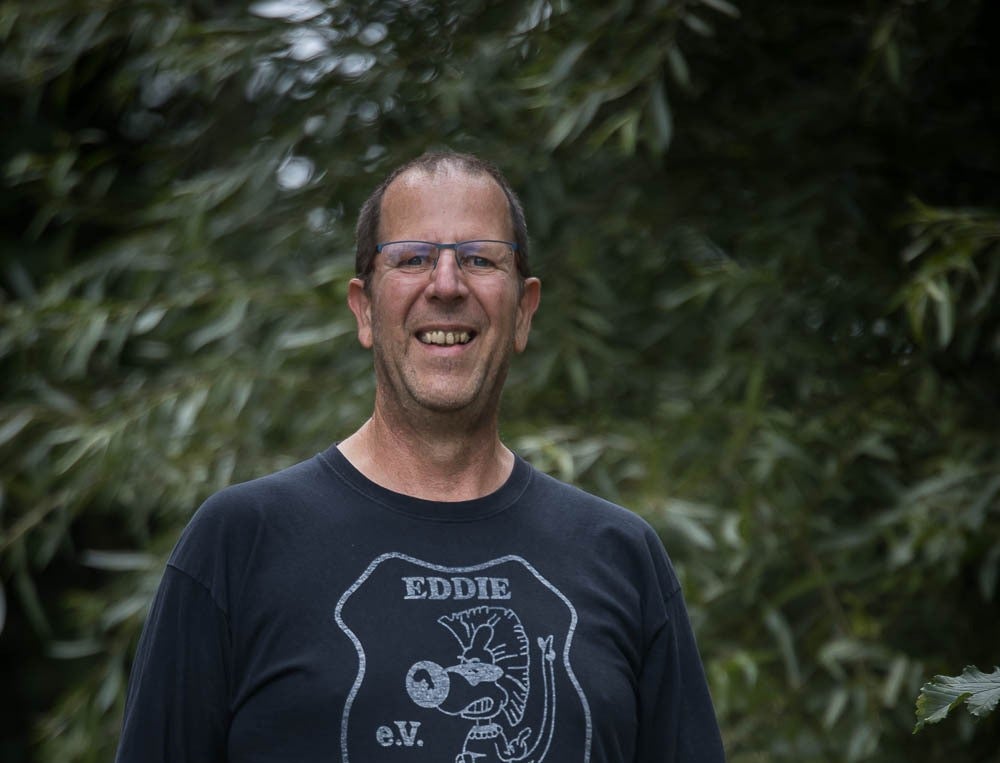 ein Mann mit schwarzem T-Shirt und Brille lacht in die Kamera. Im Hintergrund sieht man Bäume