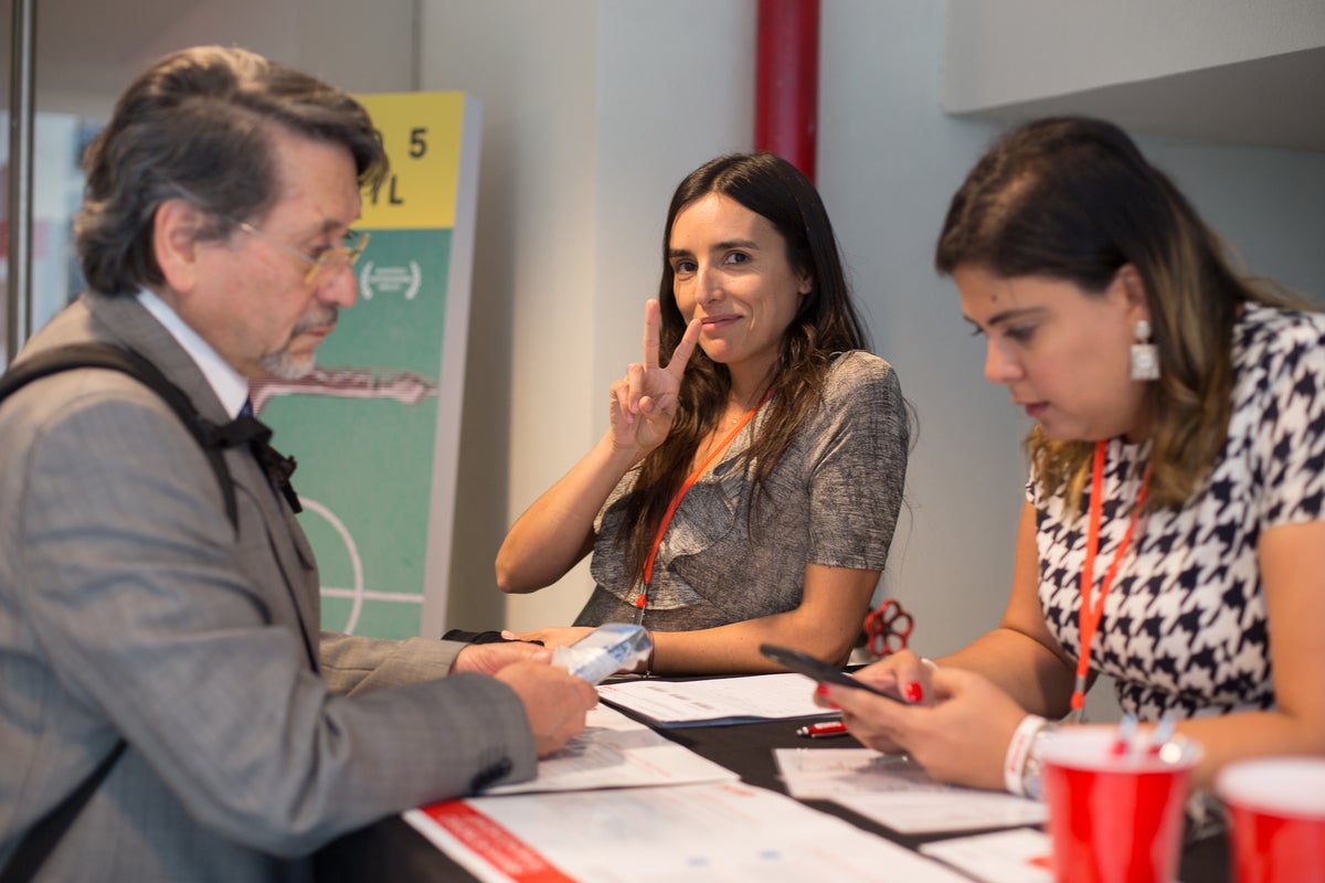 Eine Frau hält macht ein Peace Zeichen mit den Fingern, zwei andere Personen rahmen sie ein