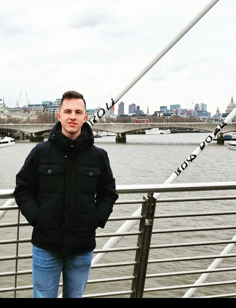  Ein Mann steht auf einer Brücke mit Metallgeländer vor einem Fluss. Er trägt eine schwarze Jacke und blaue Jeans. Im Hintergrund sind eine weitere Brücke, Boote auf dem Wasser und eine Stadtsilhouette mit modernen und historischen Gebäuden zu sehen. Der Himmel ist bewölkt.