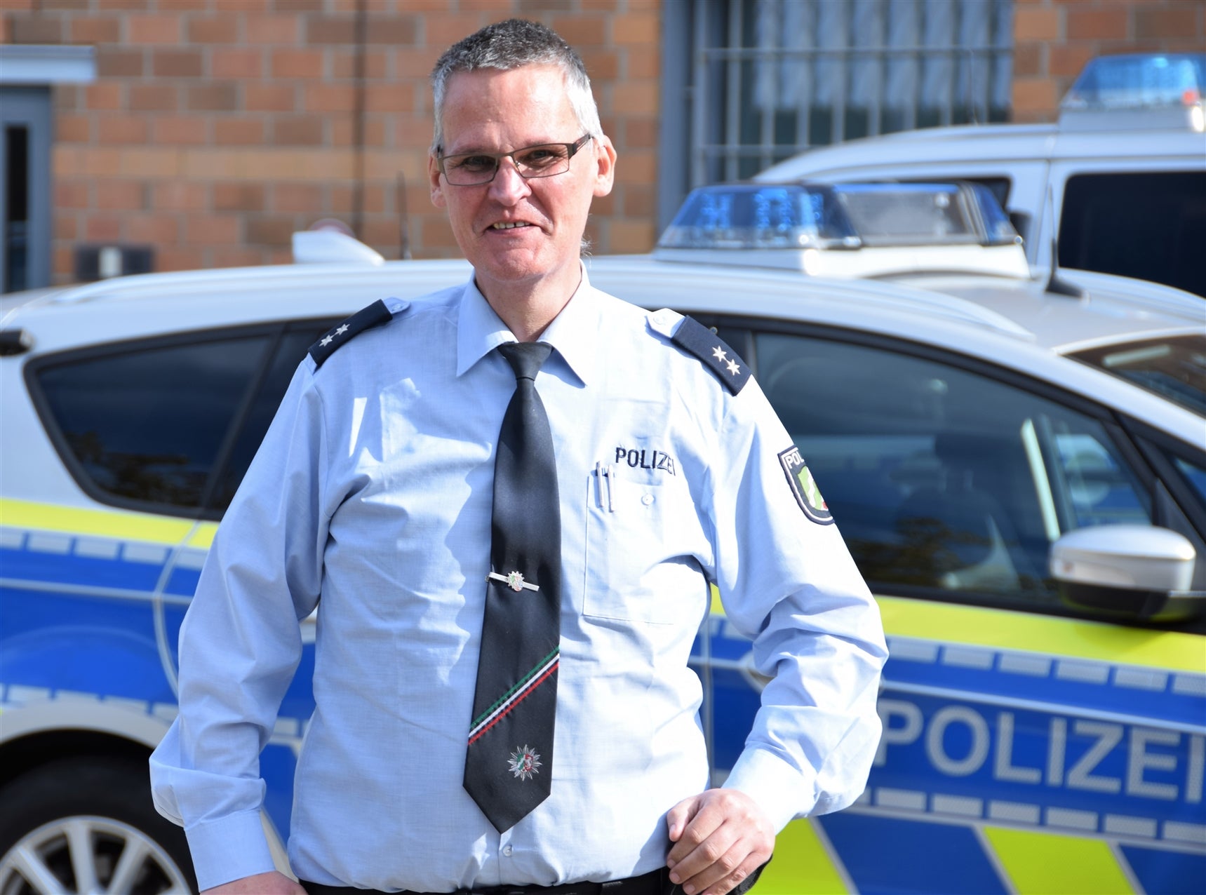 Ein Polizeibeamter in Uniform steht vor einem Polizeiwagen. Er trägt eine hellblaue Dienstbluse mit Schulterklappen, eine dunkle Krawatte mit Abzeichen sowie eine Brille. Im Hintergrund ist ein weiteres Polizeifahrzeug und ein Gebäude mit Backsteinfassade und vergitterten Fenstern zu sehen.









