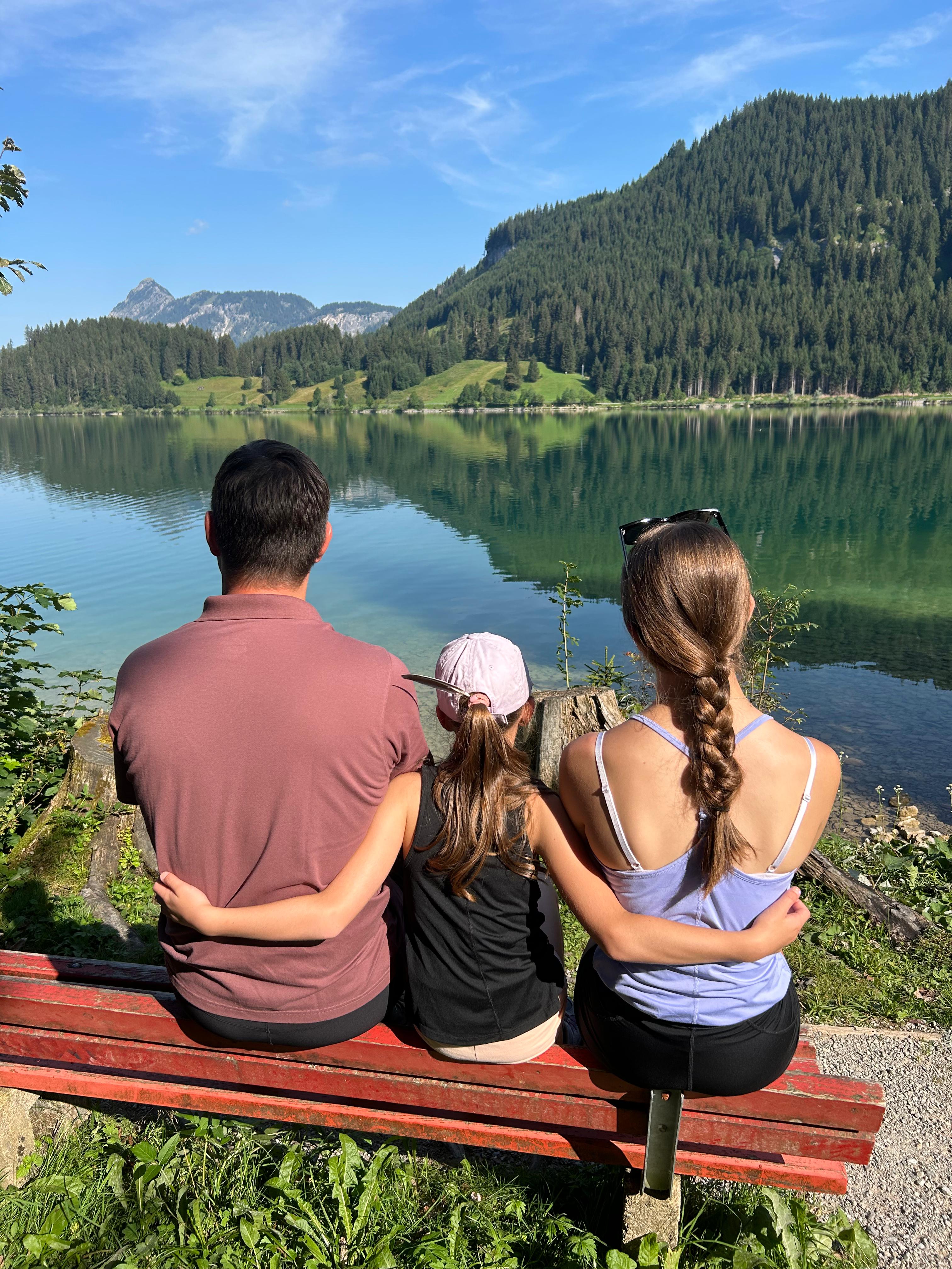 Christoph und seine Töchter sitzen auf einer Bank und schauen auf einen See.