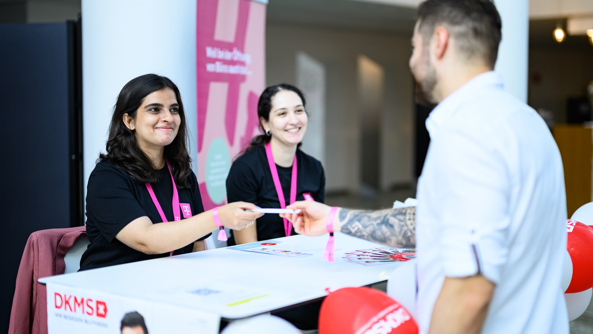 Zwei junge Frauen mit pinken DKMS-Lanyards sitzen hinter einem Infostand und lächeln. Eine von ihnen überreicht einem Mann im Vordergrund ein Formular oder Set. Der Mann steht mit dem Rücken zur Kamera. Auf dem Tisch sind DKMS-Logos, Flyer und rote Luftballons zu sehen. Im Hintergrund ist ein Banner mit dem Schriftzug „Herzlich willkommen“.








