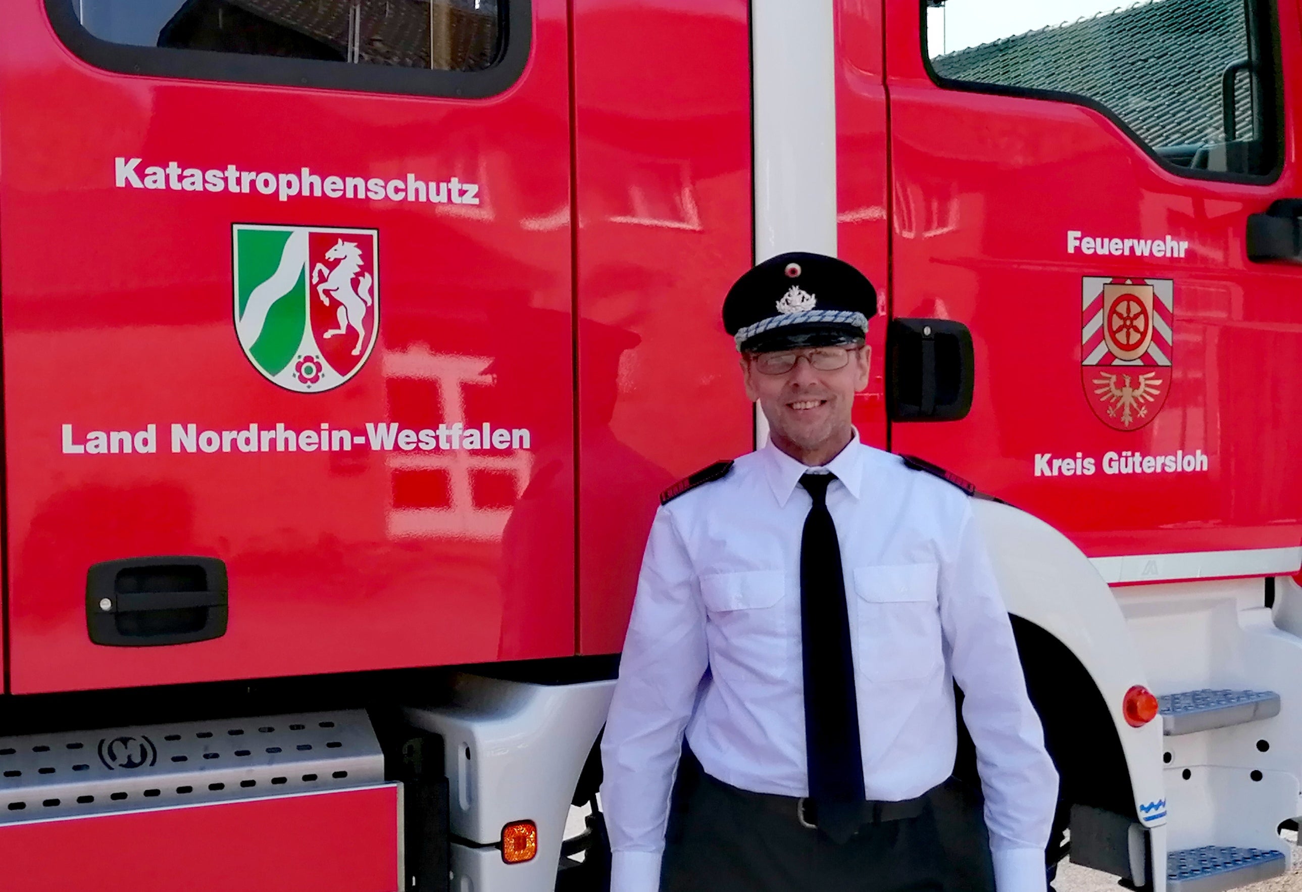 Ein Feuerwehrmann in weißem Hemd, schwarzer Krawatte und Uniformmütze steht vor einem roten Einsatzfahrzeug. Auf dem Fahrzeug steht „Katastrophenschutz Land Nordrhein-Westfalen“ sowie „Feuerwehr Kreis Gütersloh“, daneben sind die Landes- und Kreiswappen abgebildet.