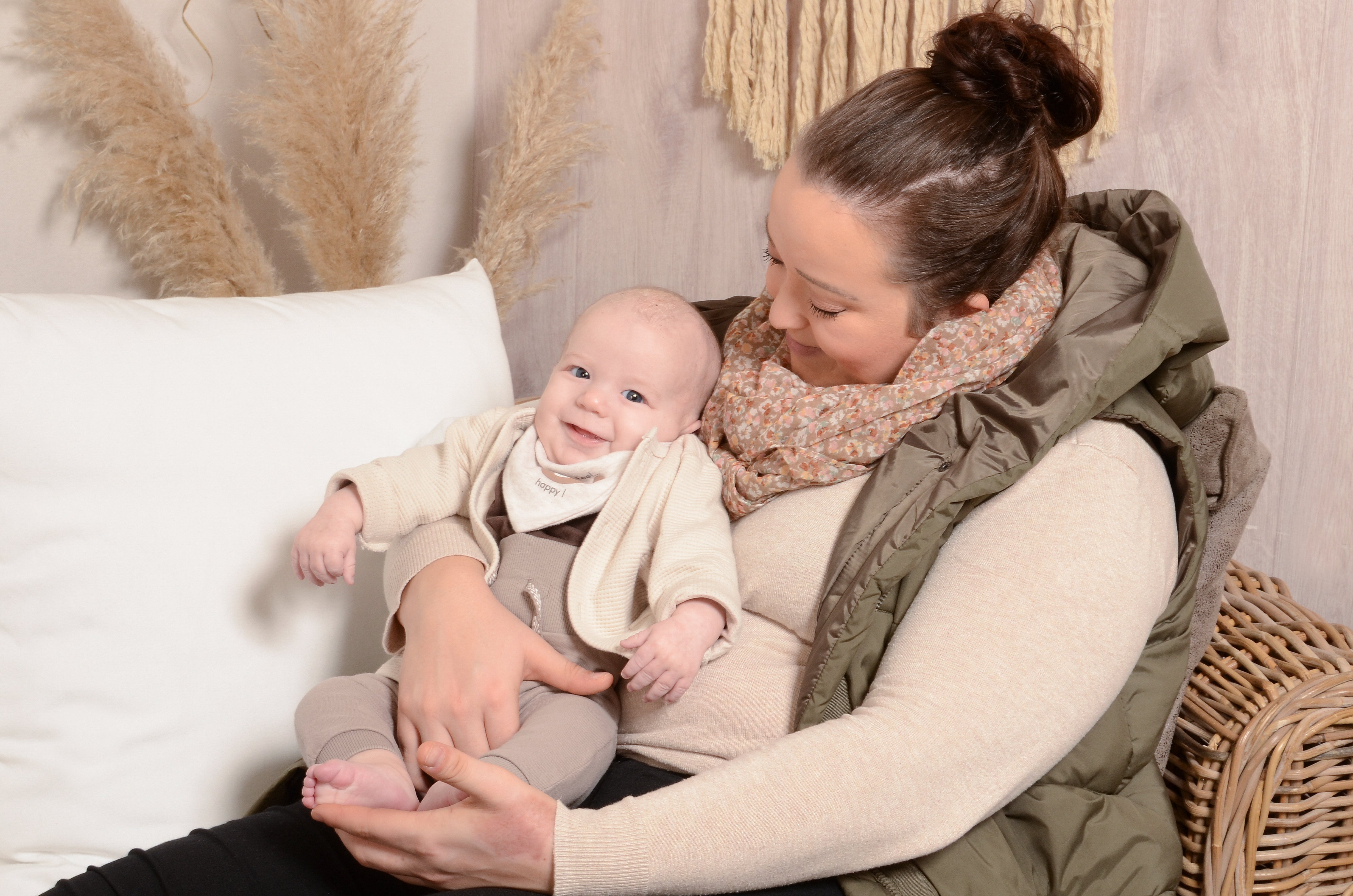 
Eine Frau sitzt auf einem Rattanstuhl und hält ein Baby im Arm. Sie schaut das Kind liebevoll an, während das Baby in die Kamera lächelt. Beide tragen helle, aufeinander abgestimmte Kleidung in Beige- und Brauntönen. Im Hintergrund sind eine weiße Kissenlehne, Pampasgras und eine Makramee-Wanddekoration zu sehen. 