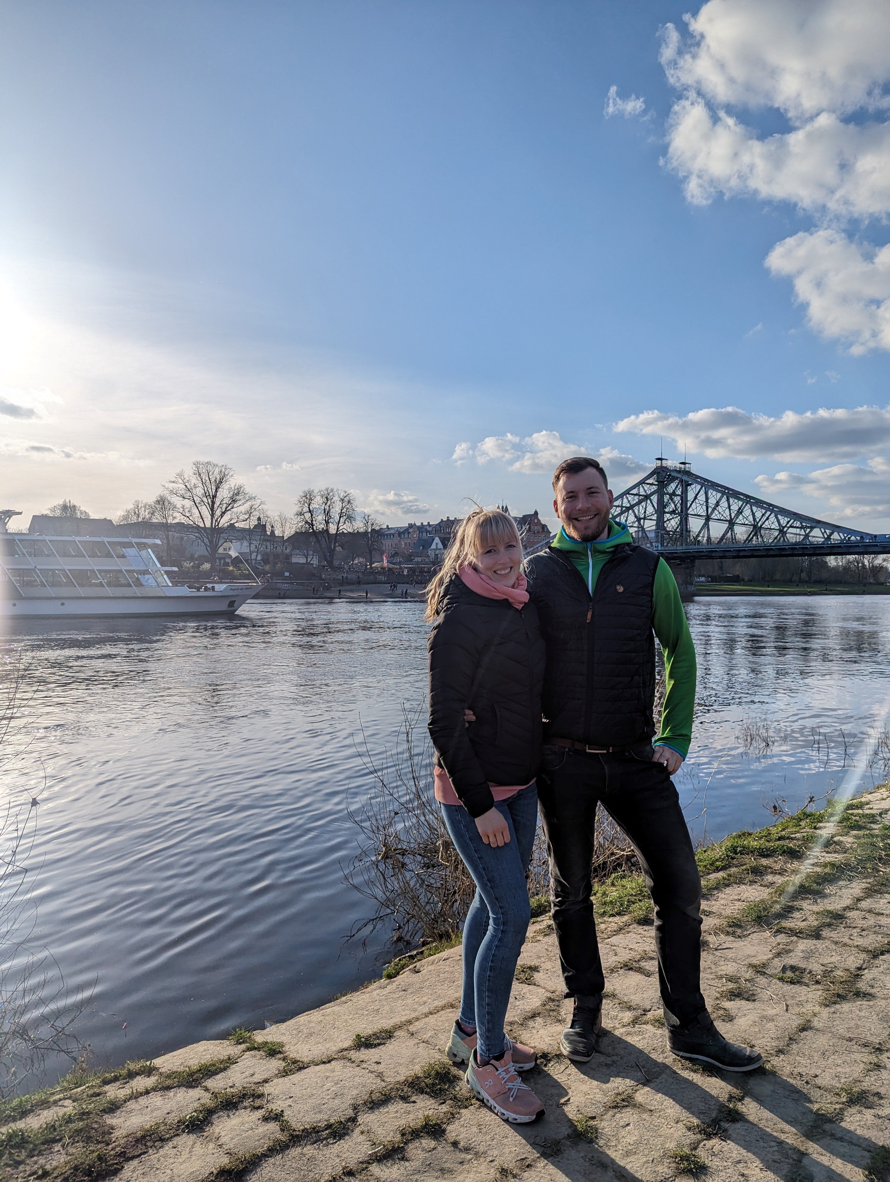 Eine Frau und ein Mann stehen an einem Flußufer, im Hintergrund sind ein Ausflugsschiff und eine Brücke zu sehen.